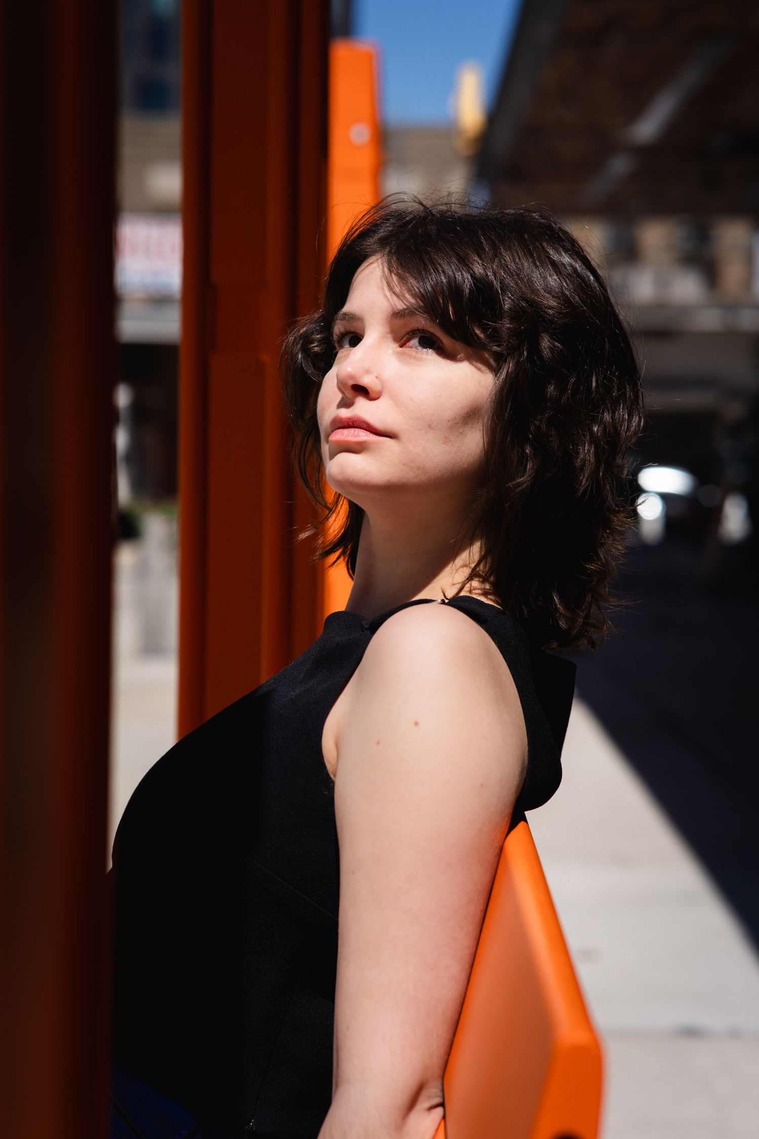 Young woman with dark hair in black sleeveless top leaning against orange pillar on sunny urban street.
