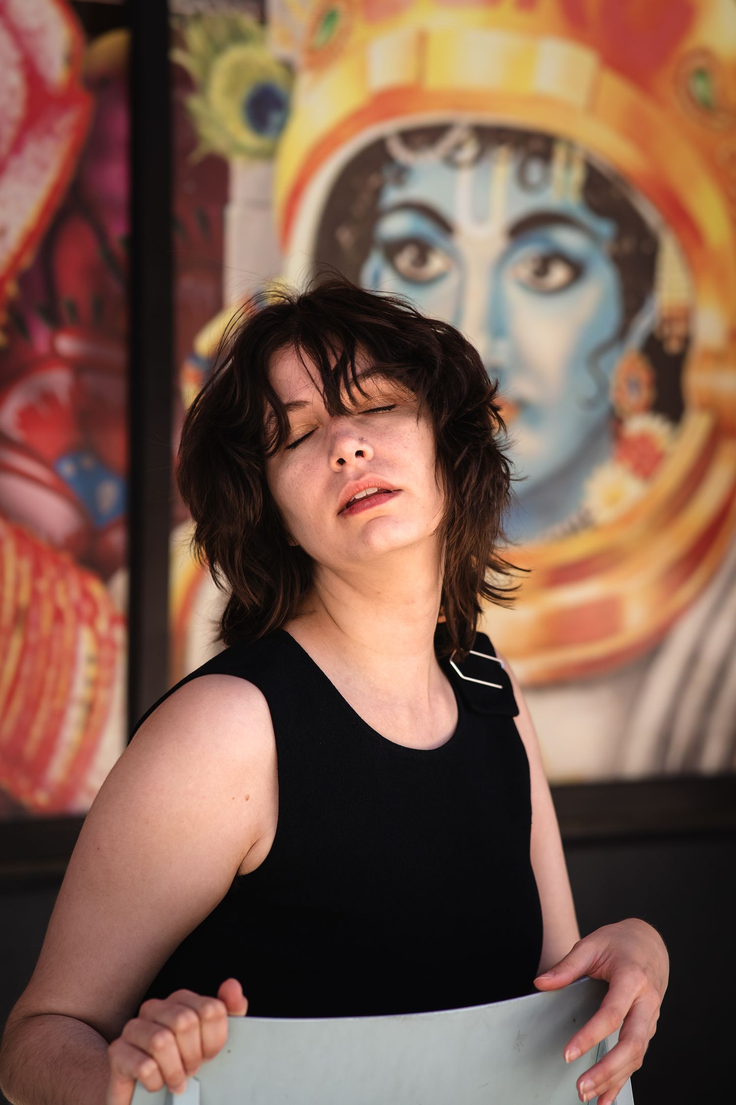 Woman in black sleeveless top poses outdoors with colorful Hindu deity mural in the background.