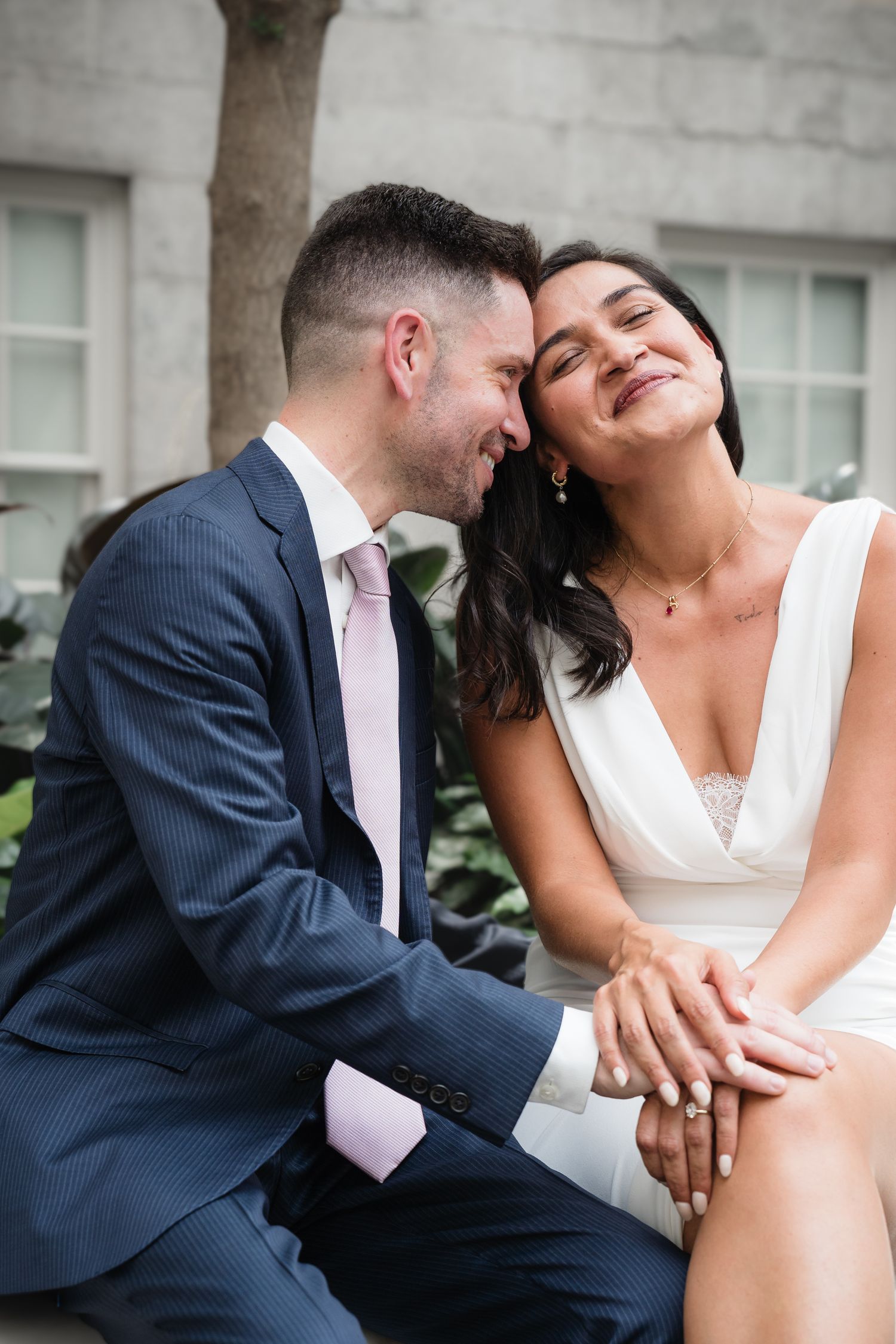 A couple in wedding attire sits closely together, smiling, as the bride displays her ring in an outdoor setting.