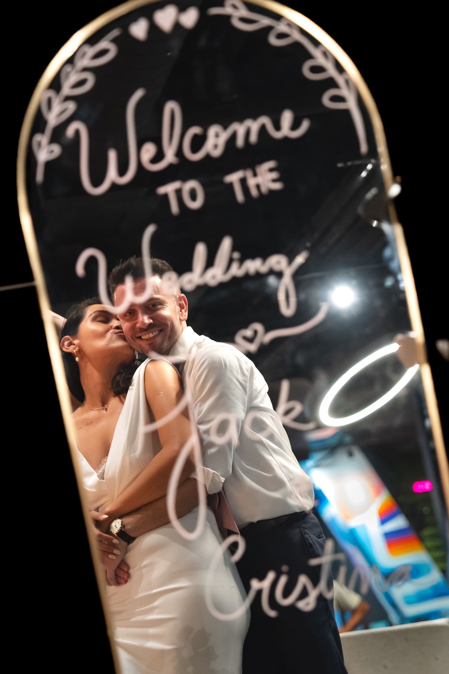 Bride and groom share a romantic kiss reflected in a wedding welcome mirror sign at their reception.