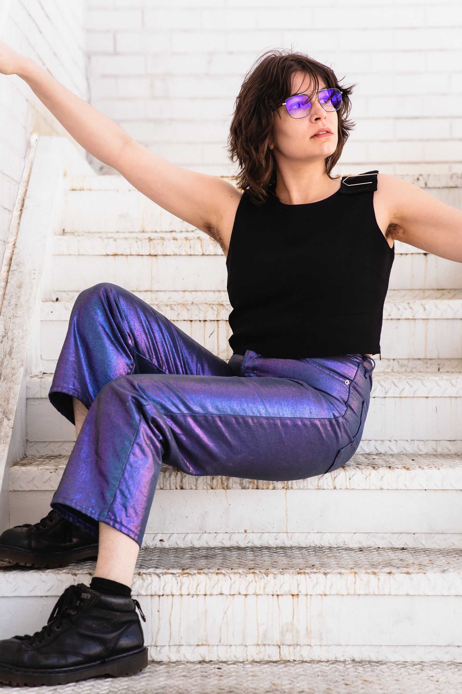 Woman in metallic purple pants, black top, purple sunglasses, and black boots sitting on white stairs.