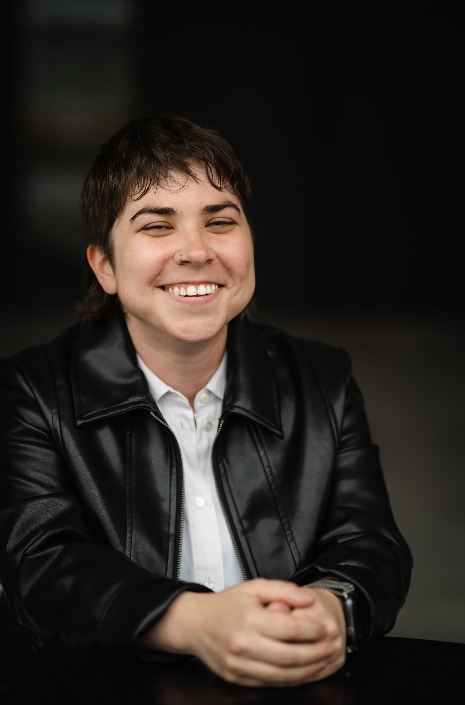 Person in black leather jacket and white shirt smiles warmly in dark setting.