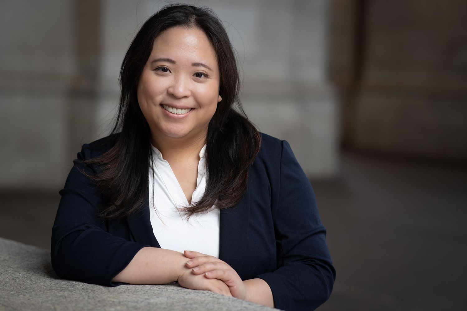 Professional woman in navy blazer smiling confidently, posing in a modern office setting for a corporate headshot.