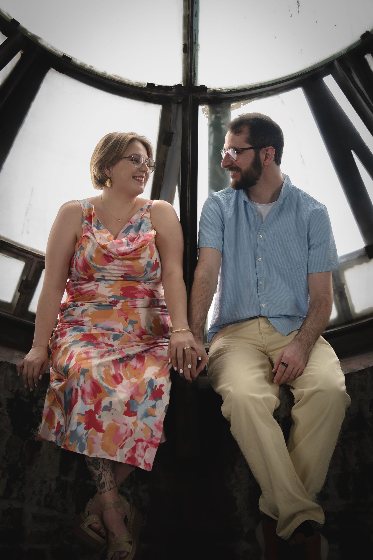 A couple in casual attire sitting together by a large window with clock mechanics.