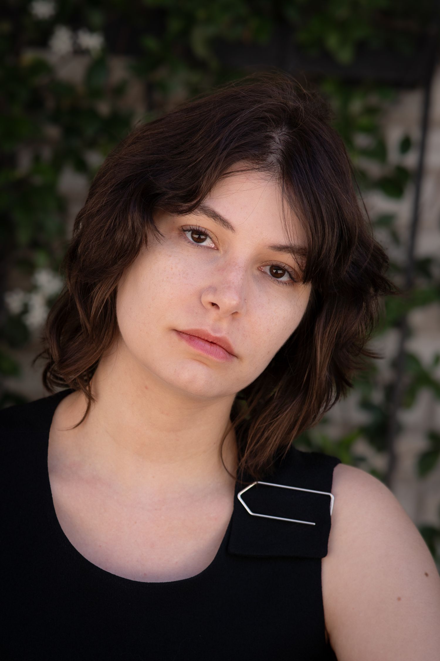Young woman with short dark wavy hair wearing a black sleeveless top, posing outdoors with green foliage background.