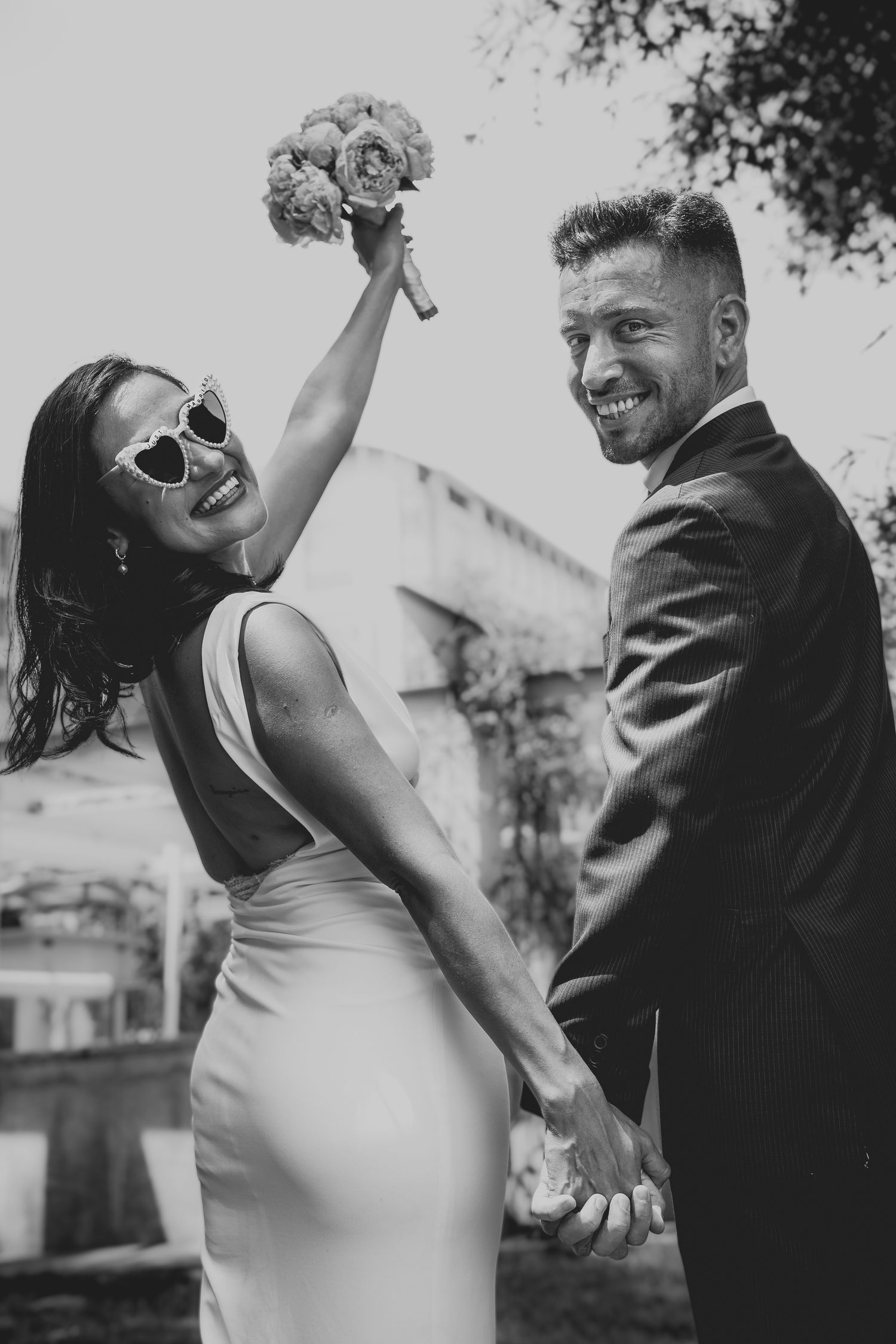 Black and white photo of a joyful bride in sunglasses raising bouquet while groom smiles holding hands outdoors.