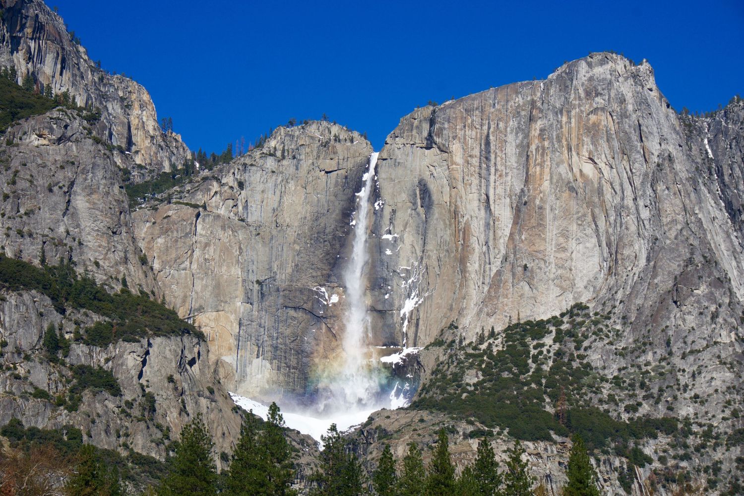 Massive granite cliffs of Yosemite Valley with dramatic waterfall under blue sky.