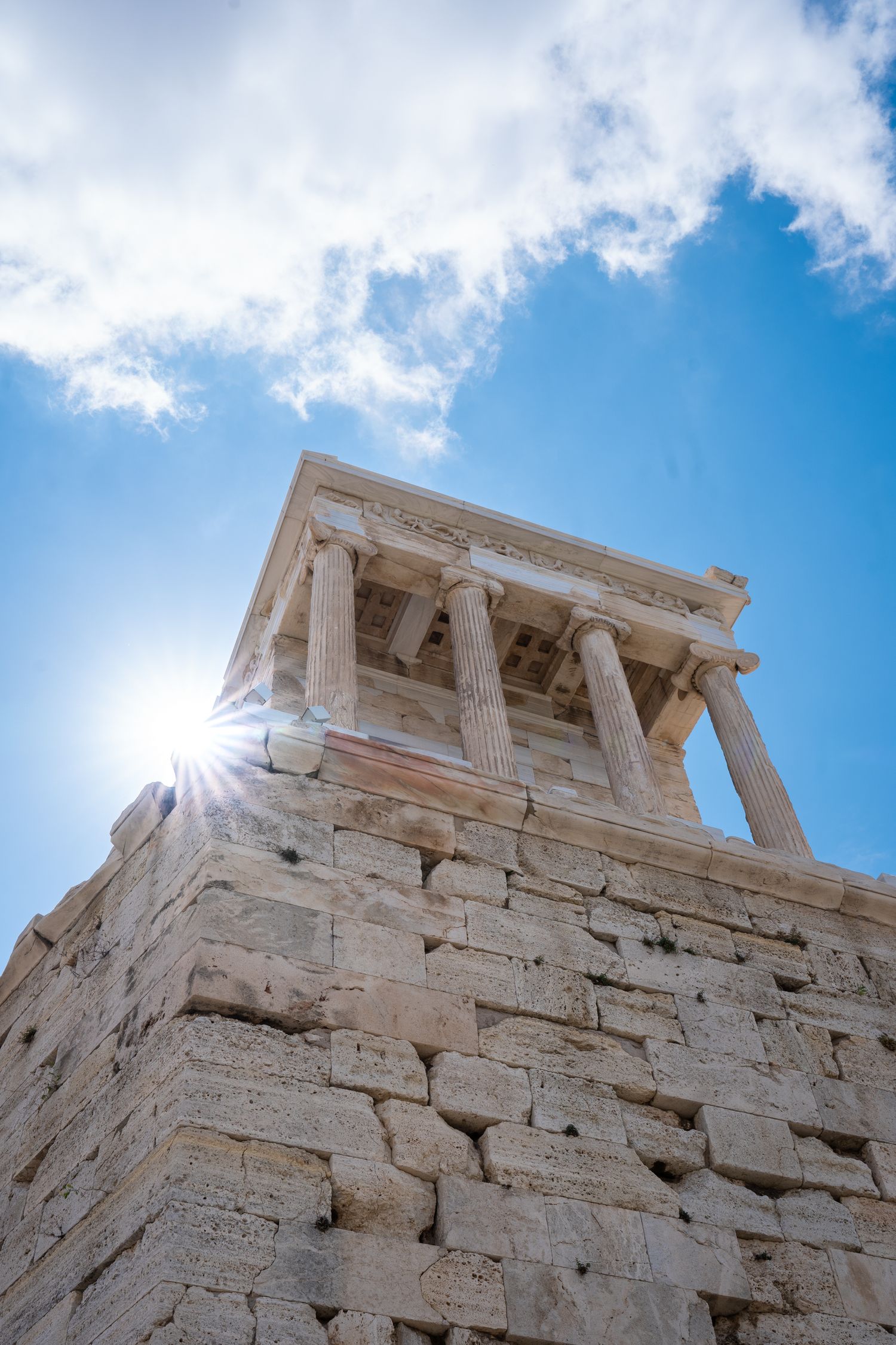 Ancient Greek temple columns and stone walls of the Acropolis rise against a bright blue sky with sunburst in Athens, Greece.