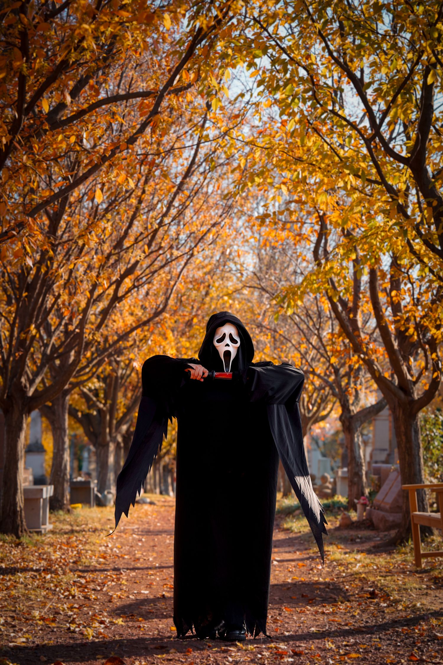 Ghostface figure standing among fall foliage in cemetery with dramatic lighting.