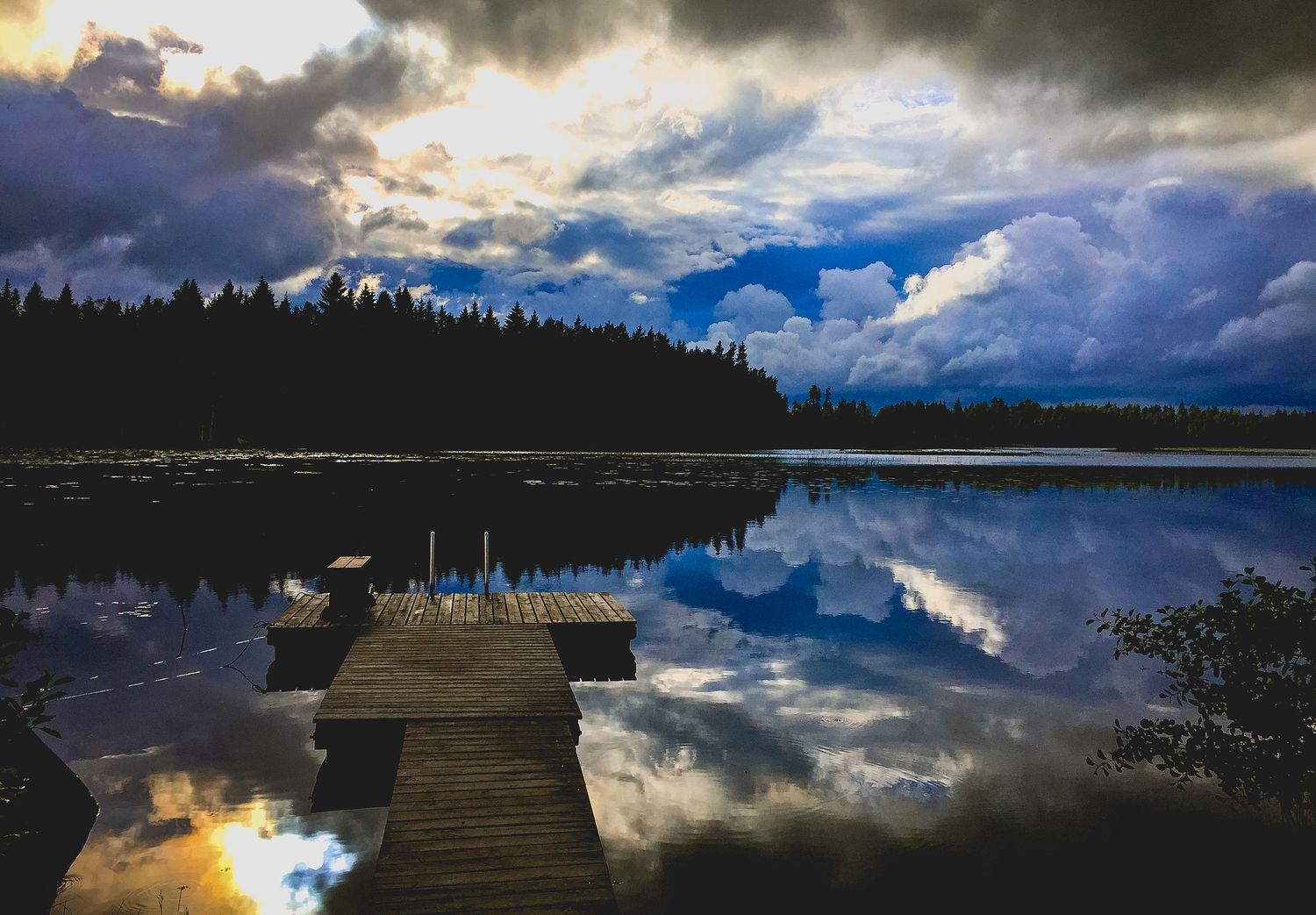 Dramatic storm clouds reflecting on calm lake water with wooden dock in foreground.