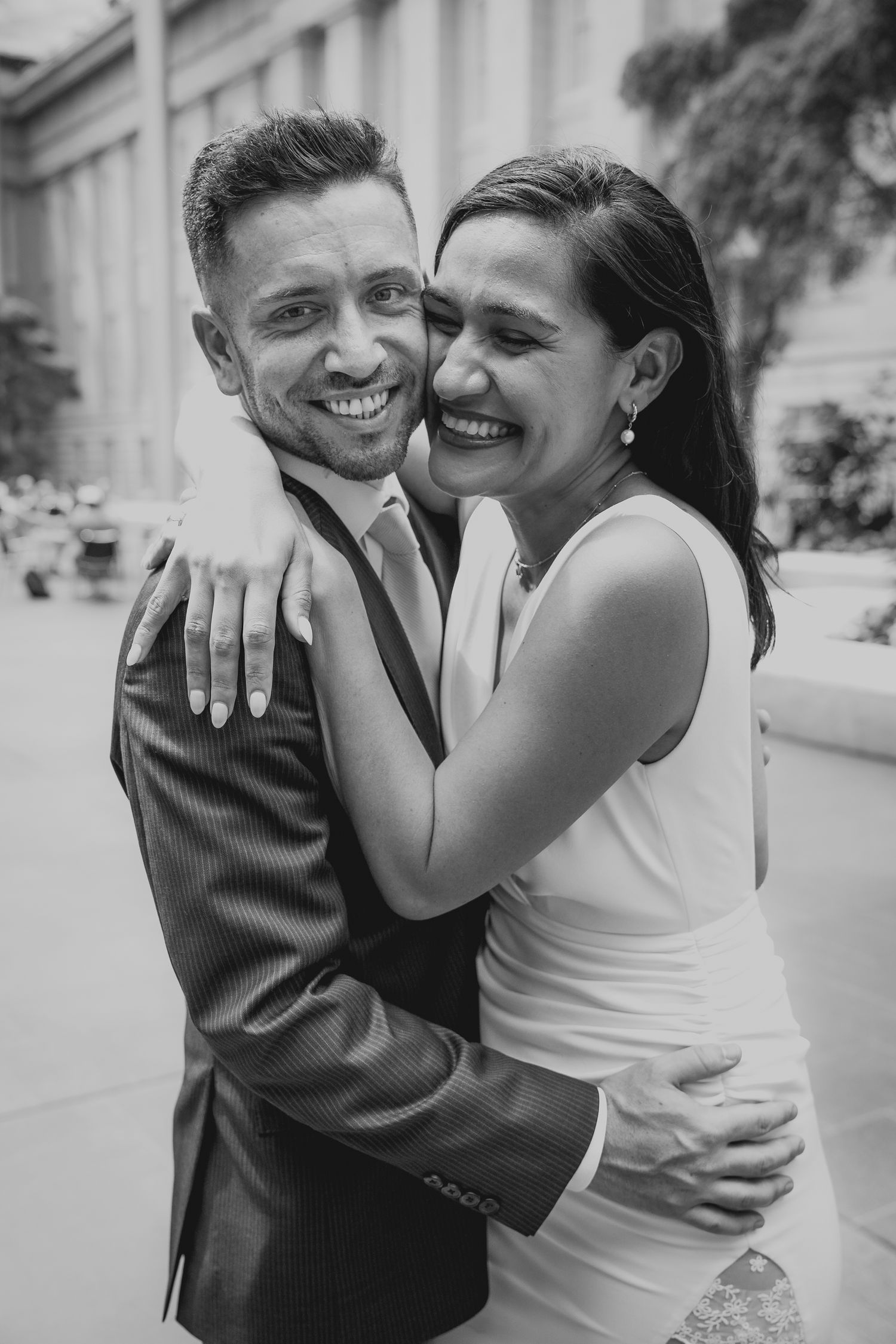 Black and white photo of joyful newlyweds embracing and laughing outside a classic stone building.