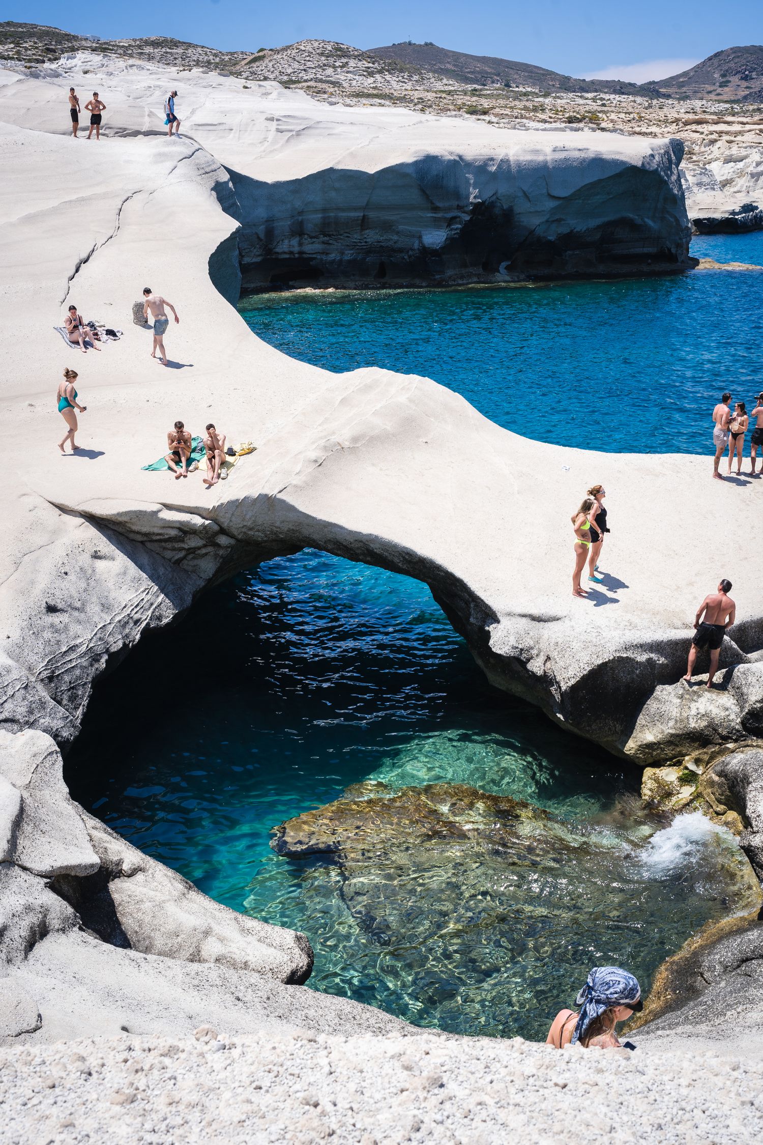 Natural rock formation with turquoise waters and swimmers at Sarakiniko Beach in Milos, Greece.
