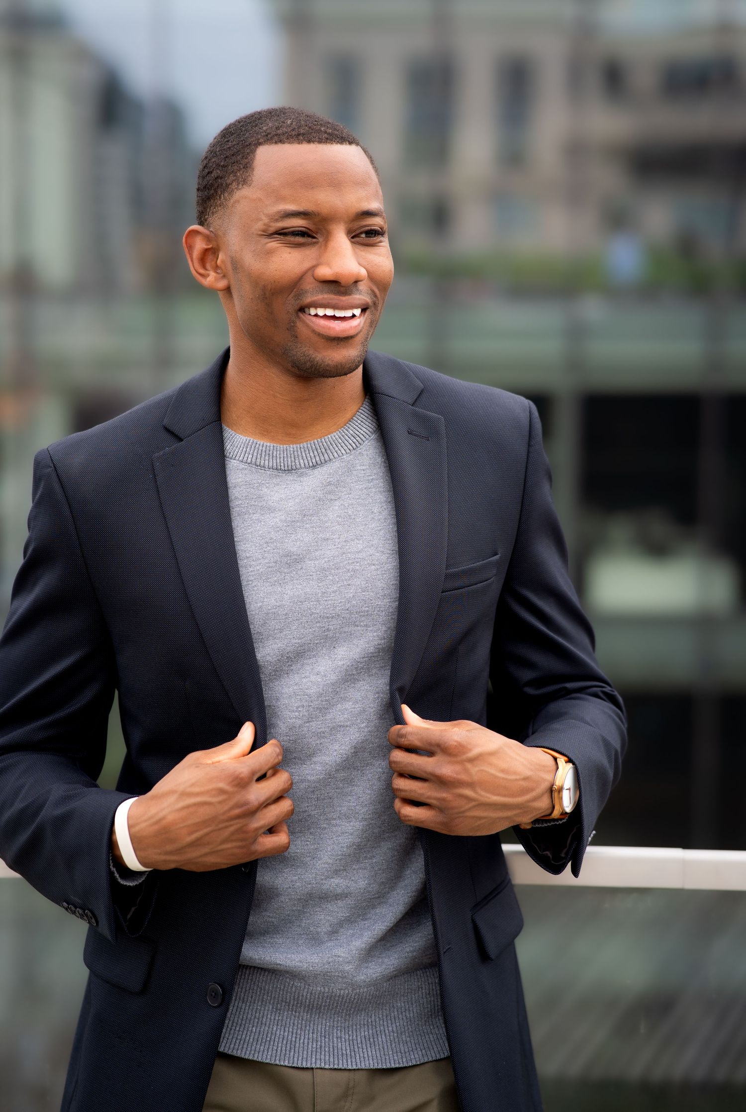 Professional wearing navy blazer and grey sweater adjusts jacket while smiling outdoors.