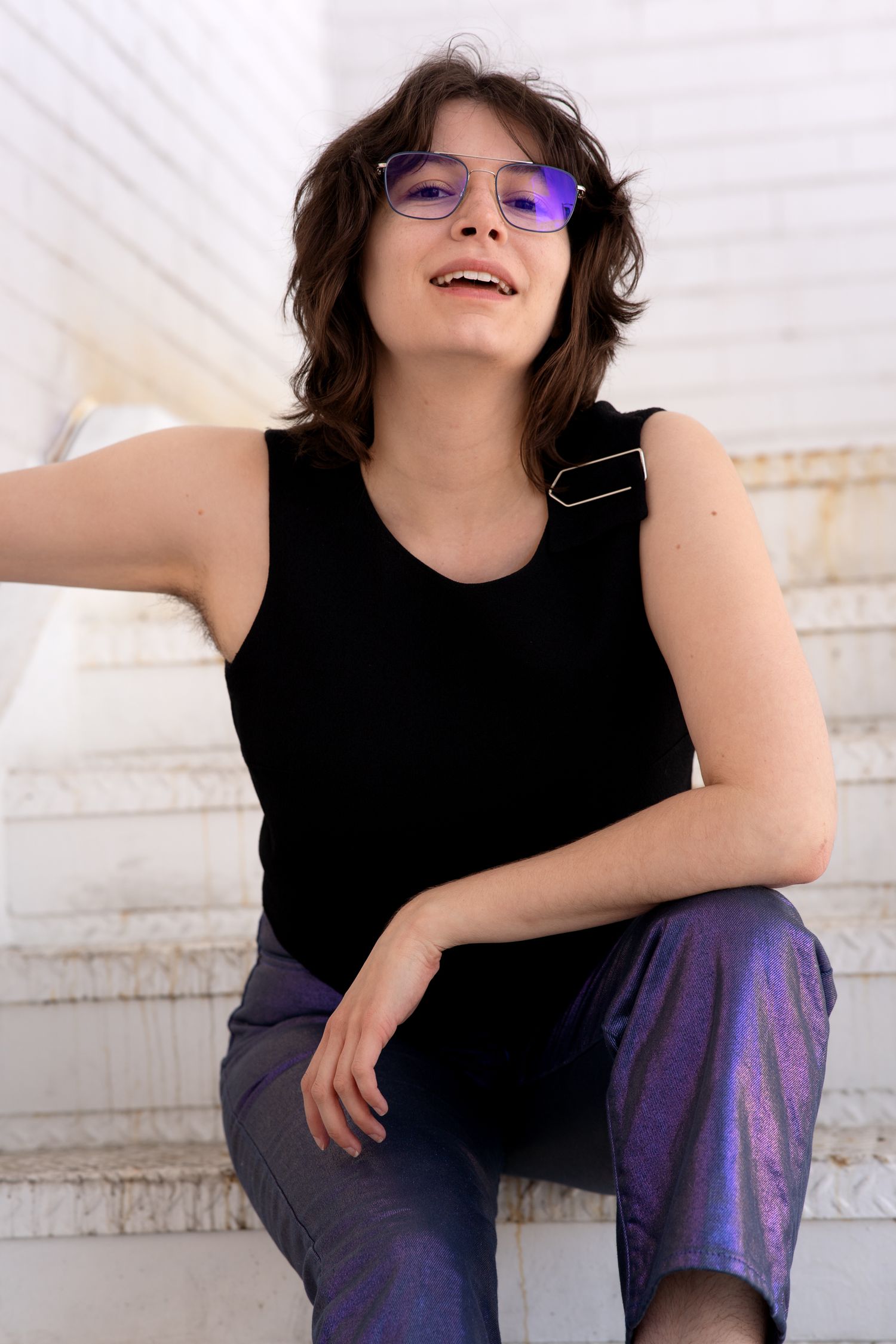 Young woman with purple glasses and black tank top sitting on white marble steps, smiling outdoors.