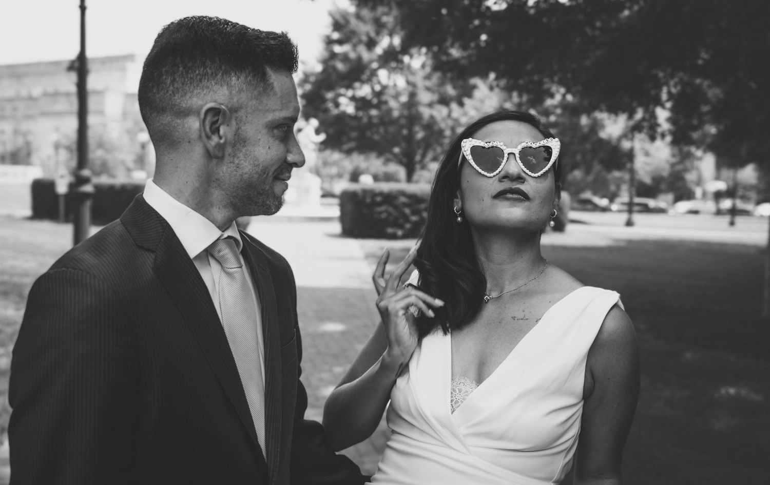 Black and white photo of a bride in sunglasses and groom in suit sharing a romantic outdoor moment.