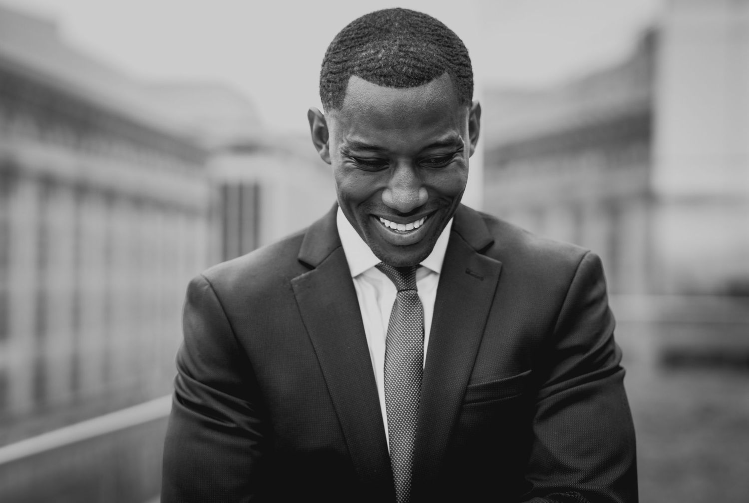Professional in black suit and tie smiling warmly against urban backdrop in black and white portrait.
