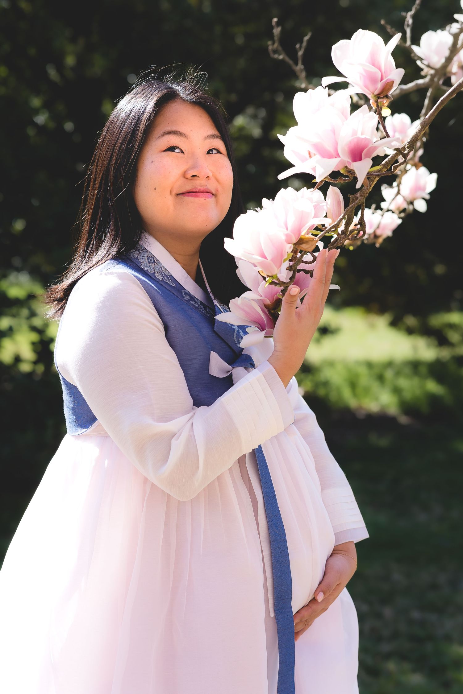 Woman in traditional Korean dress admiring pink magnolia blossoms in garden.