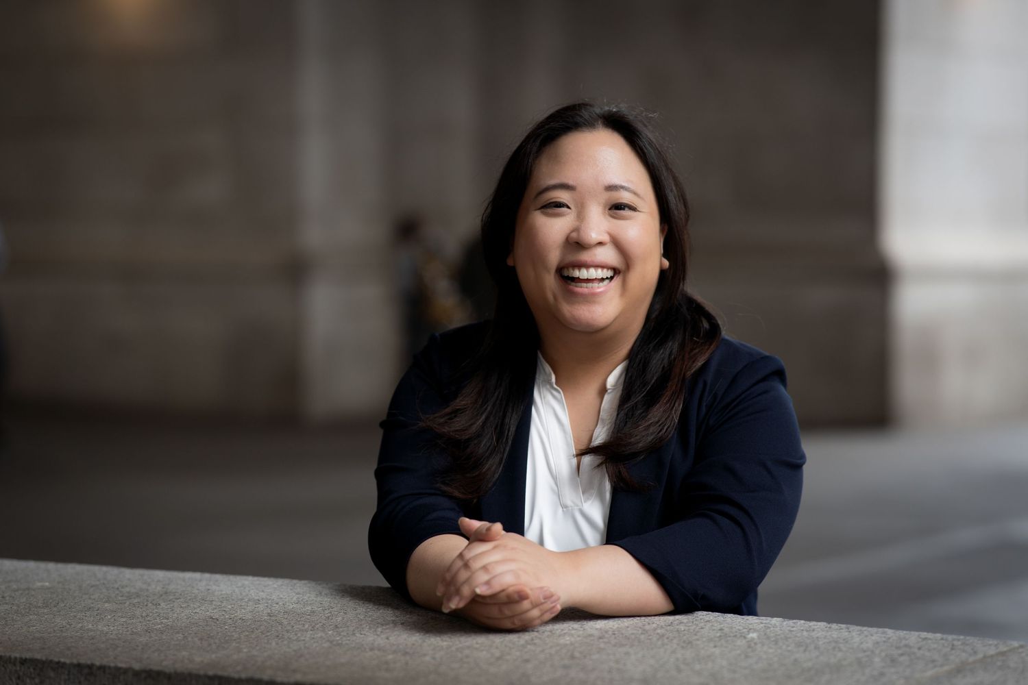 Smiling Asian woman in navy blazer leaning on stone ledge in modern architectural setting.