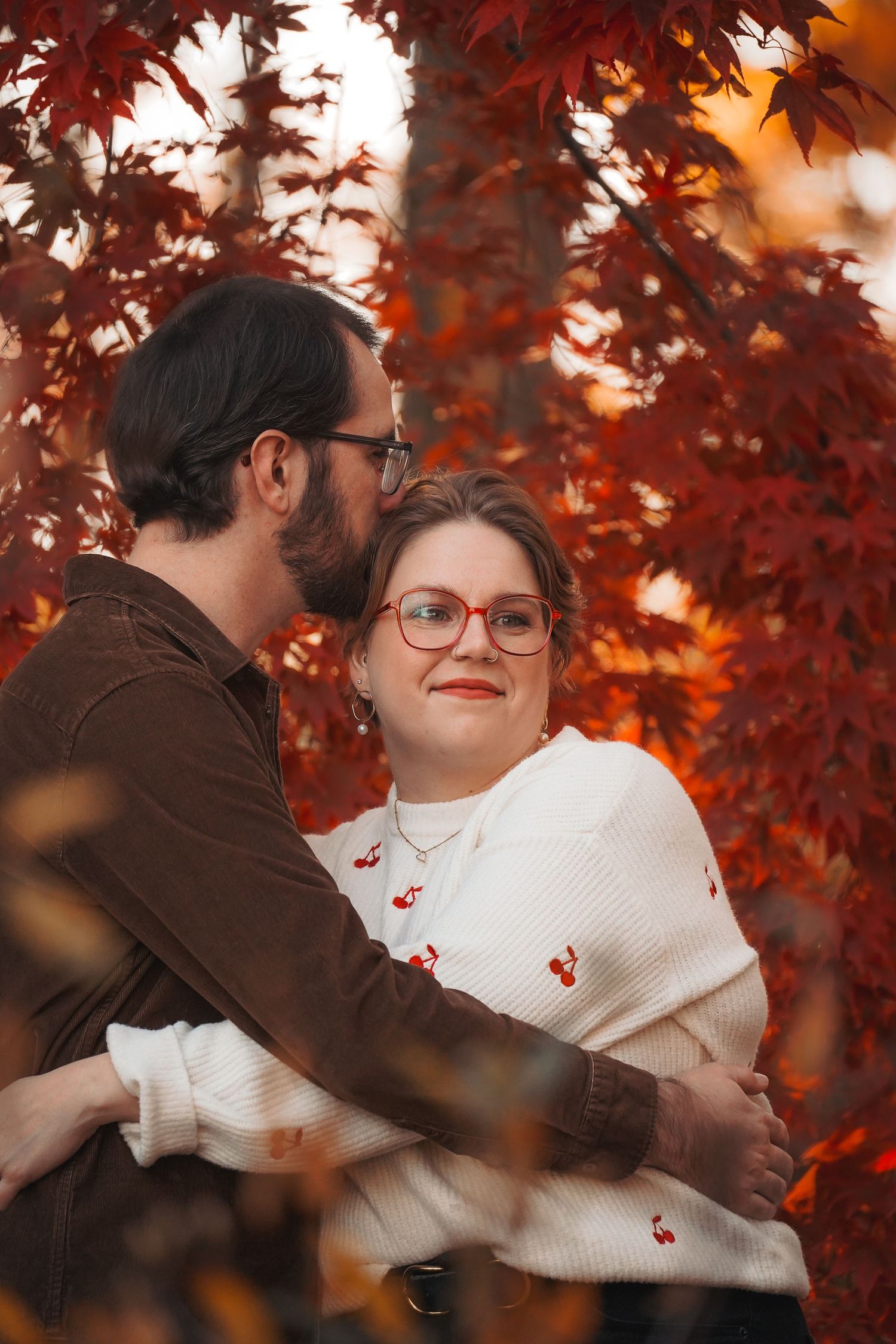 A couple embraces under vibrant red autumn maple leaves in a romantic outdoor setting.