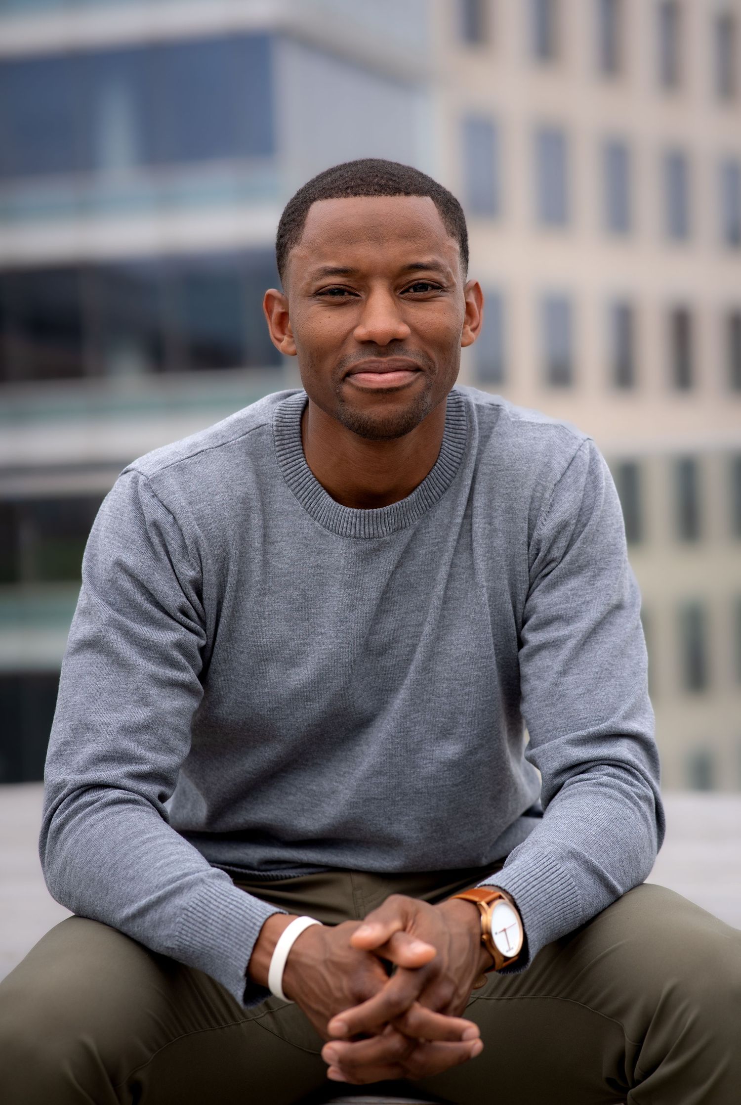Young professional in grey sweater sitting casually outdoors with city buildings in background.