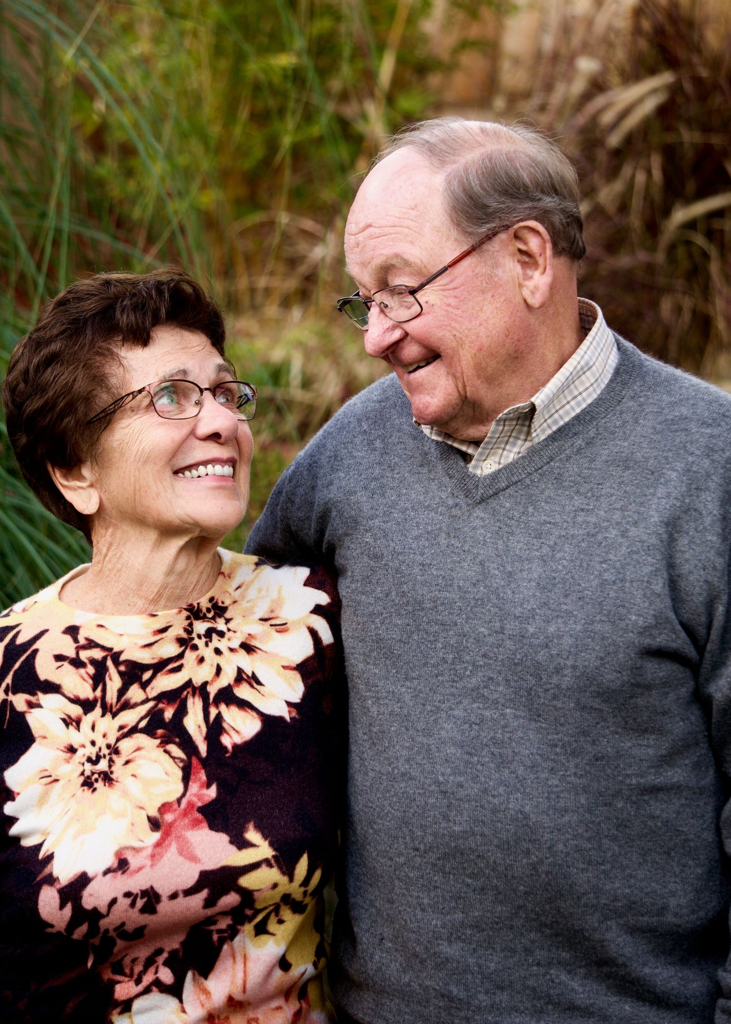 An elderly couple shares an intimate moment while smiling at each other outdoors.