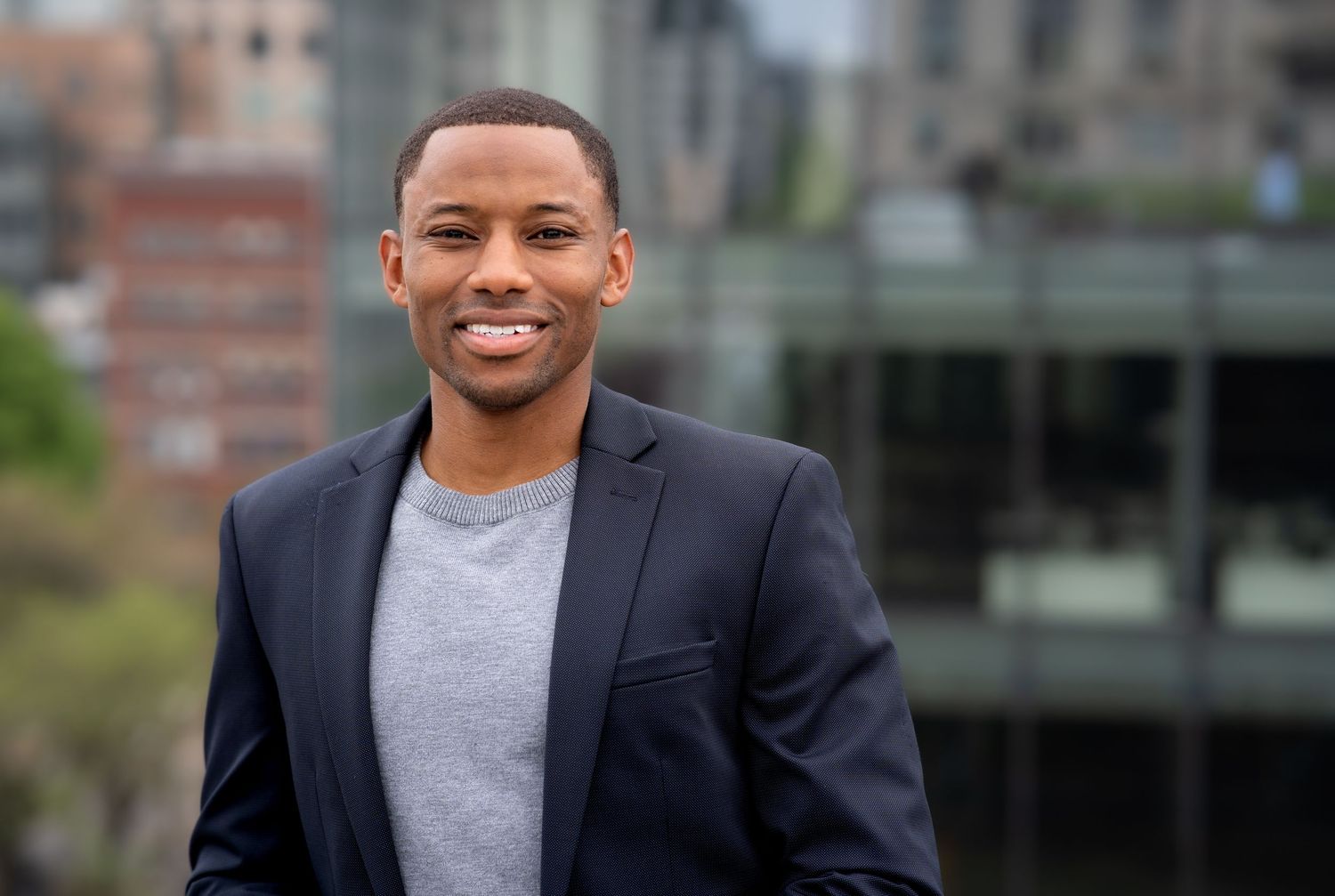 Professional in dark blazer and grey shirt poses confidently against urban background.