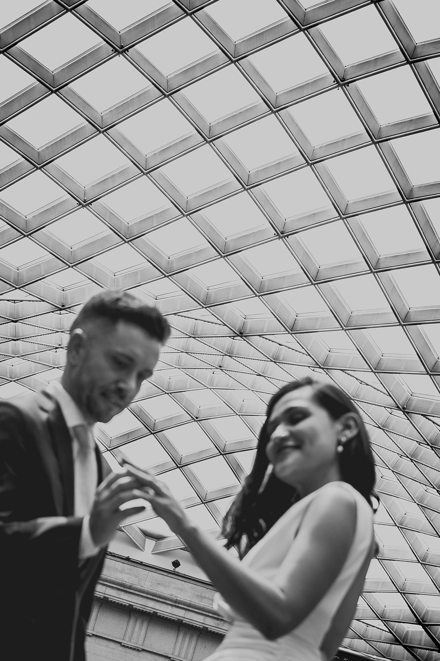 Black and white photo of a smiling couple looking at a phone under a geometric glass ceiling.