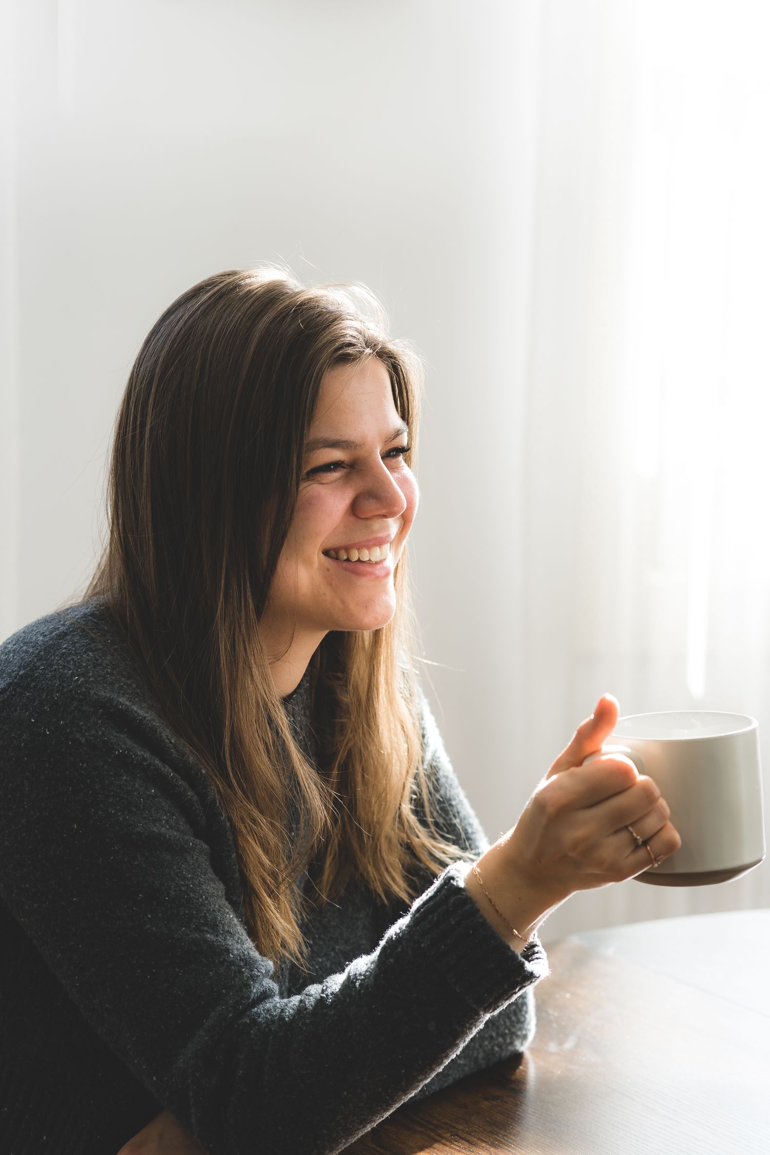 Woman in dark sweater smiling while holding coffee cup near window.