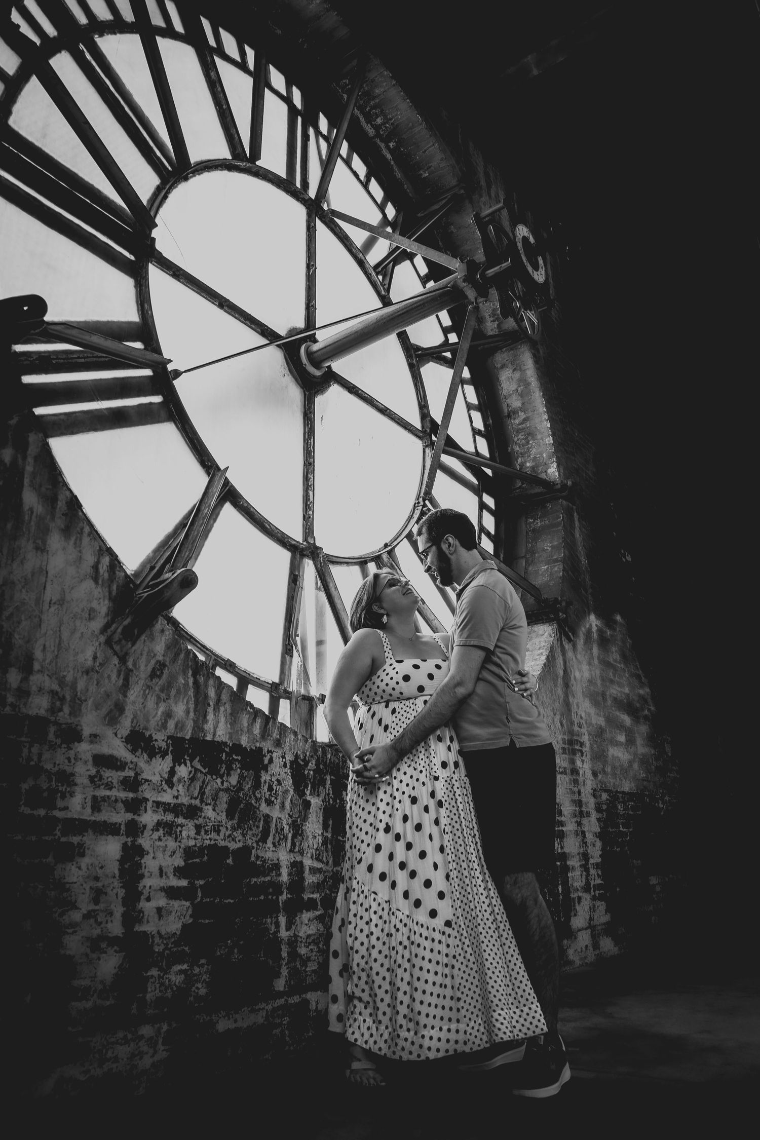 Black and white photo of a couple posing by a large clock face.