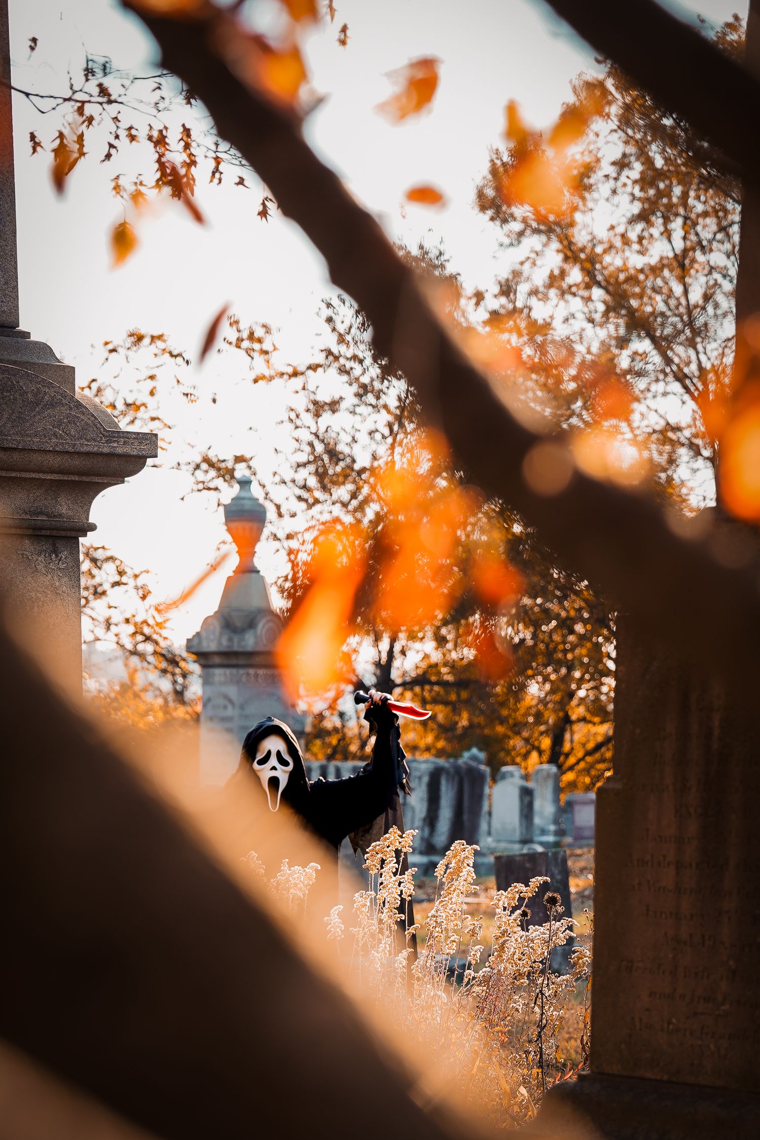 Ghostface mask visible through autumn leaves in cemetery setting.