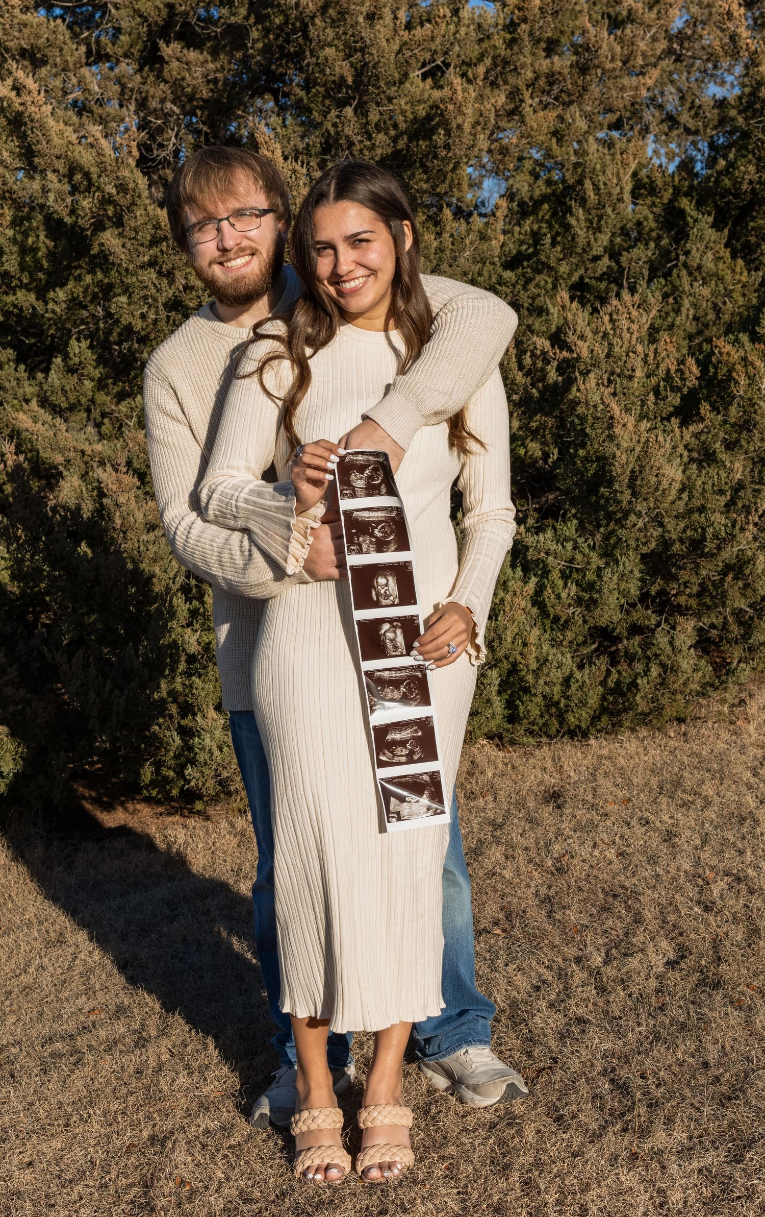 Mom and dad holding ultrasound pictures for baby announcement in beautiful outdoor lighting.
