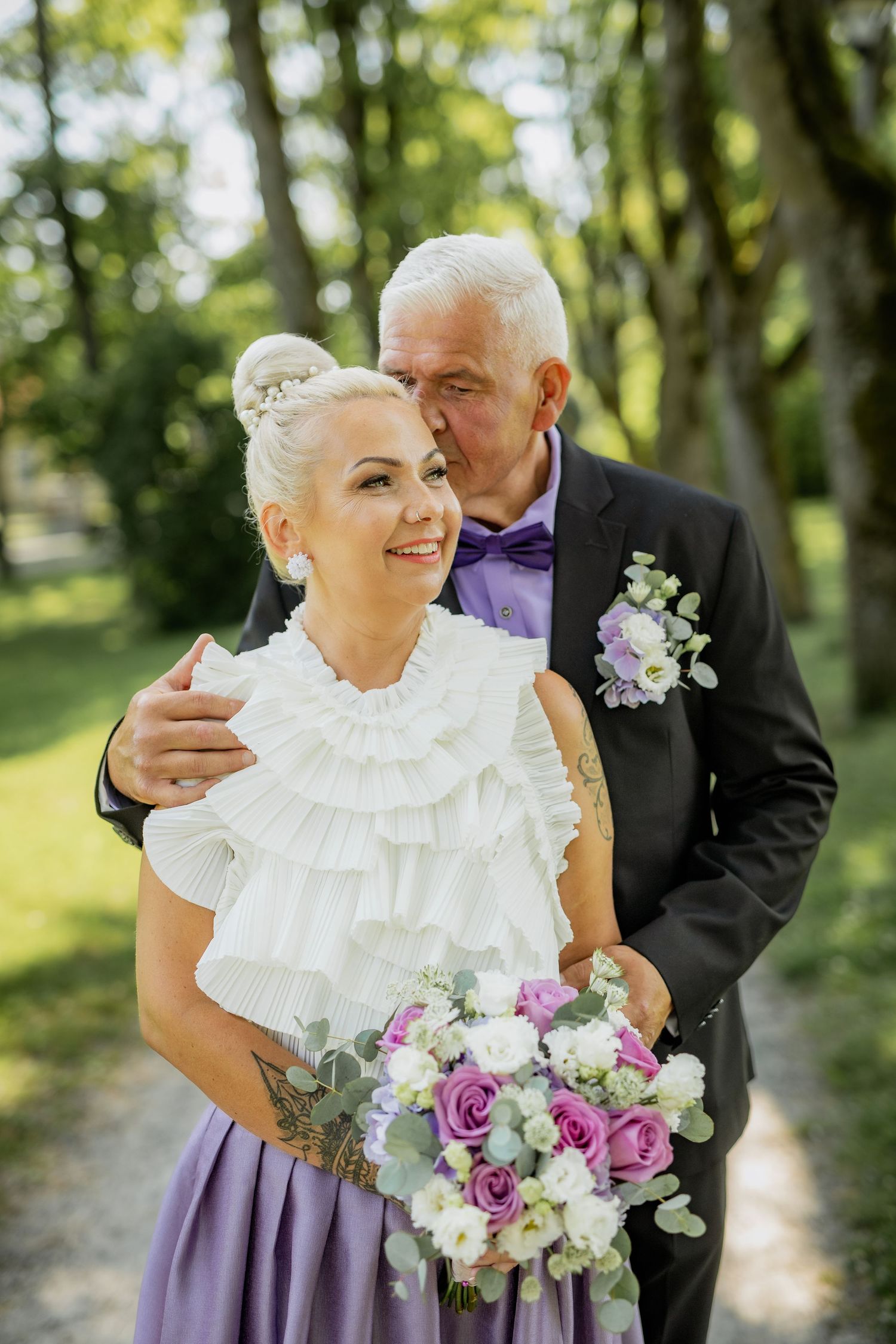 Older groom kisses bride's temple as she smiles holding purple and white bouquet in a tree-lined park.