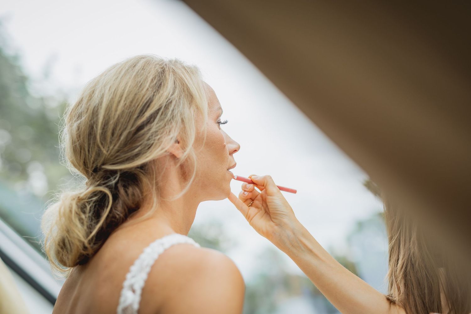 Makeup artist applying lipstick to a blonde bride with an elegant updo before her wedding ceremony.