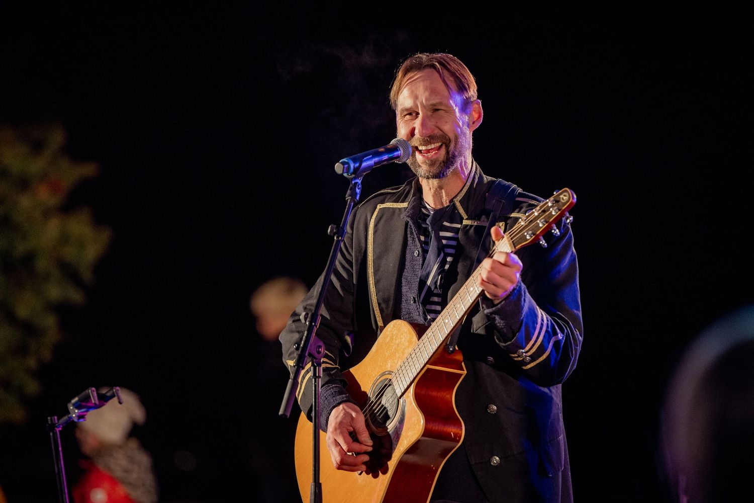 Smiling male guitarist performing live on stage at night, playing acoustic guitar in a dark navy jacket.