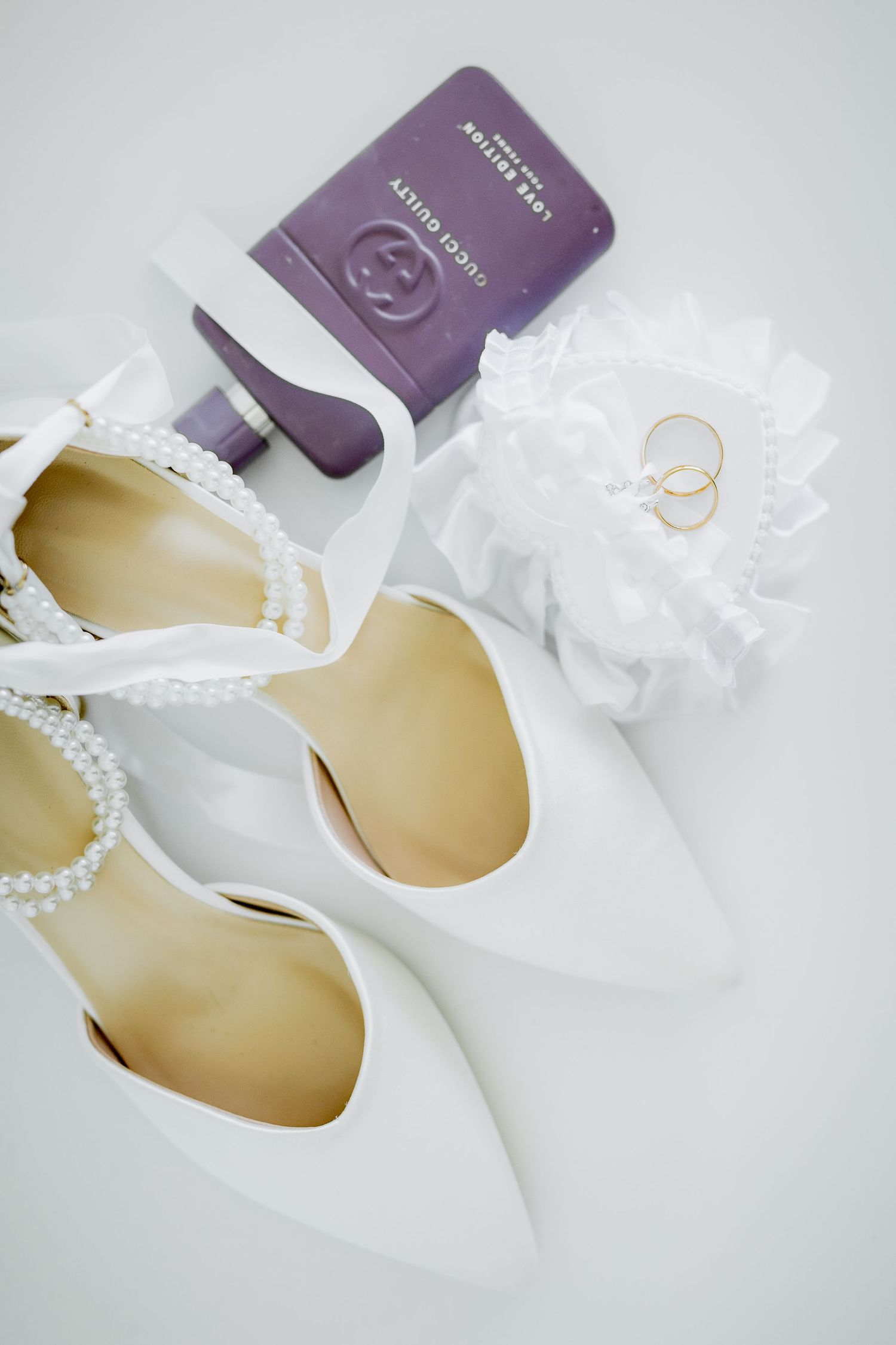 White pearl-strap bridal heels with Gucci perfume, gold wedding rings on lace garter on white background.