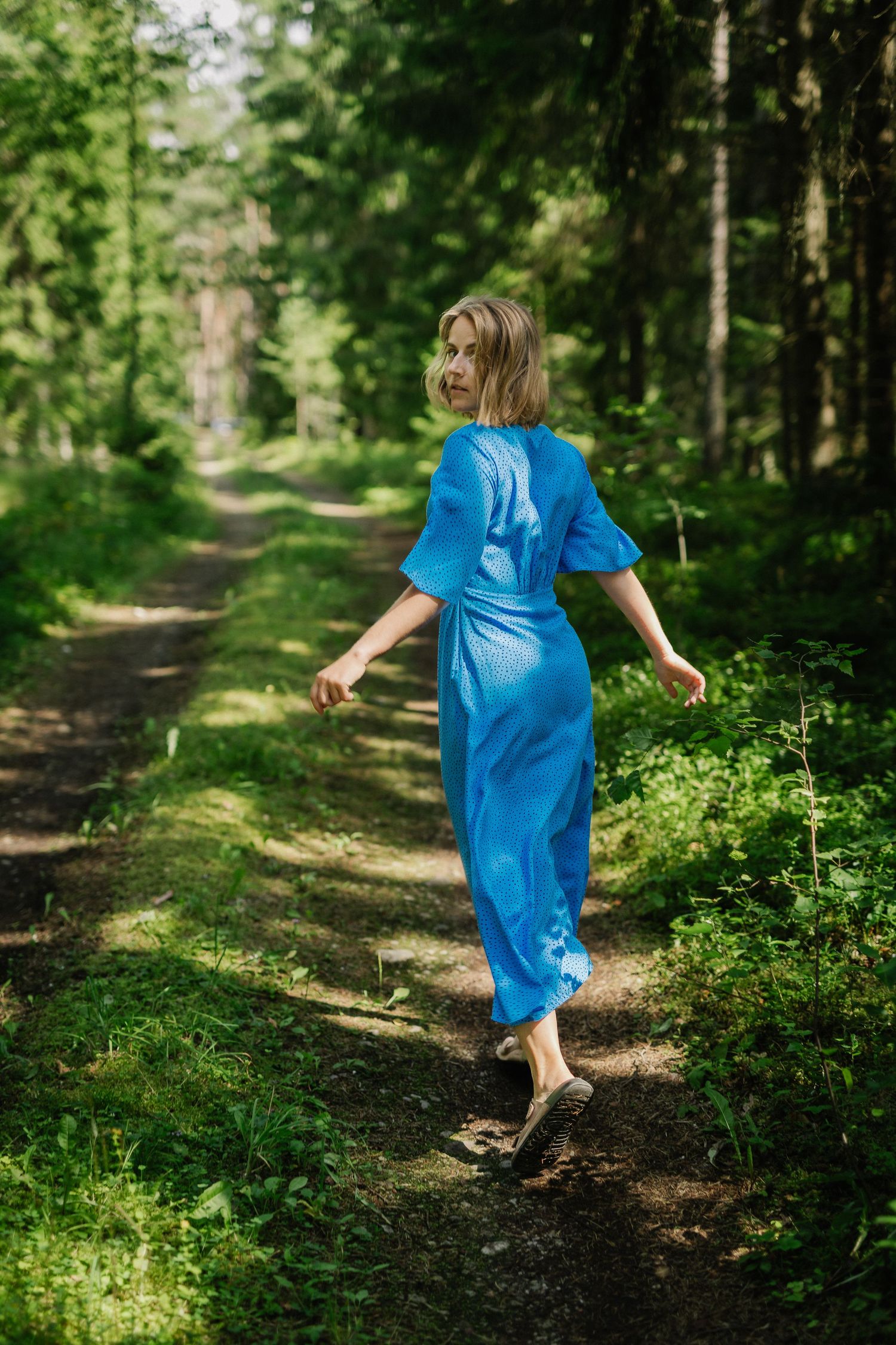 Woman in blue midi dress walks barefoot along green forest path, glancing back over shoulder.