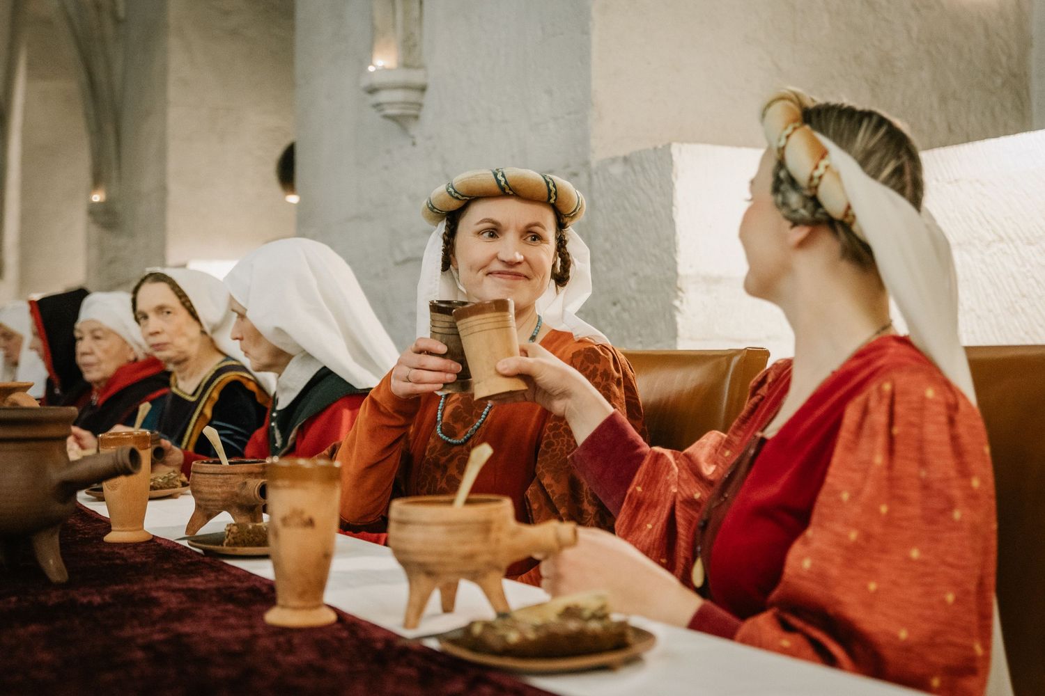 Women in medieval costumes dining together, sharing drinks and food in a historic castle setting.
