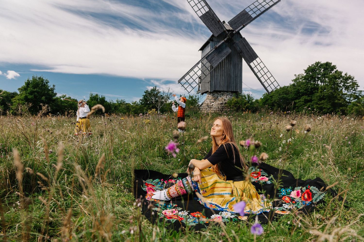 Woman in traditional dress sits in wildflower meadow with windmill and people in background on cloudy day.