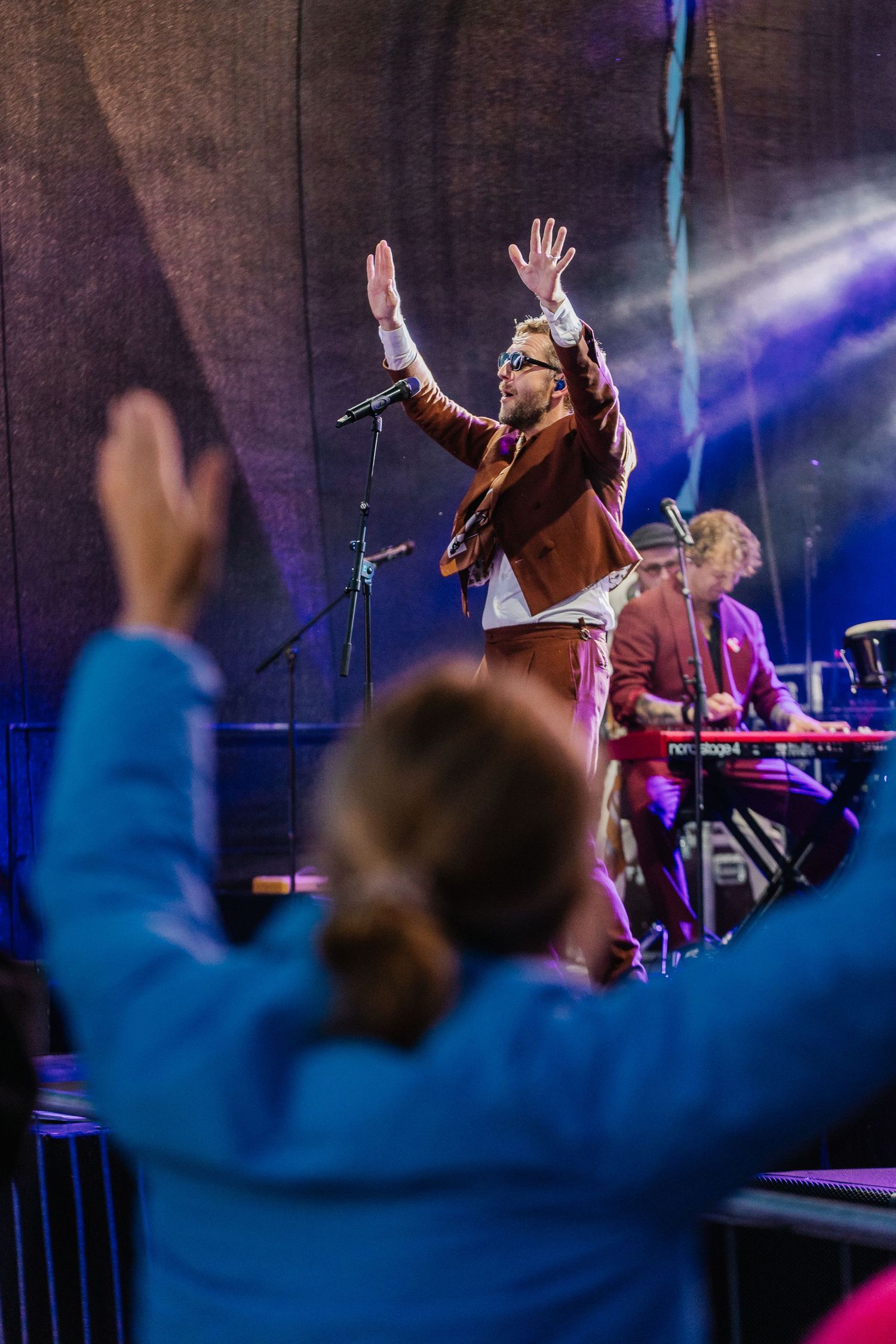 Performer in brown jacket raises hands on stage while audience members cheer at an outdoor music festival.
