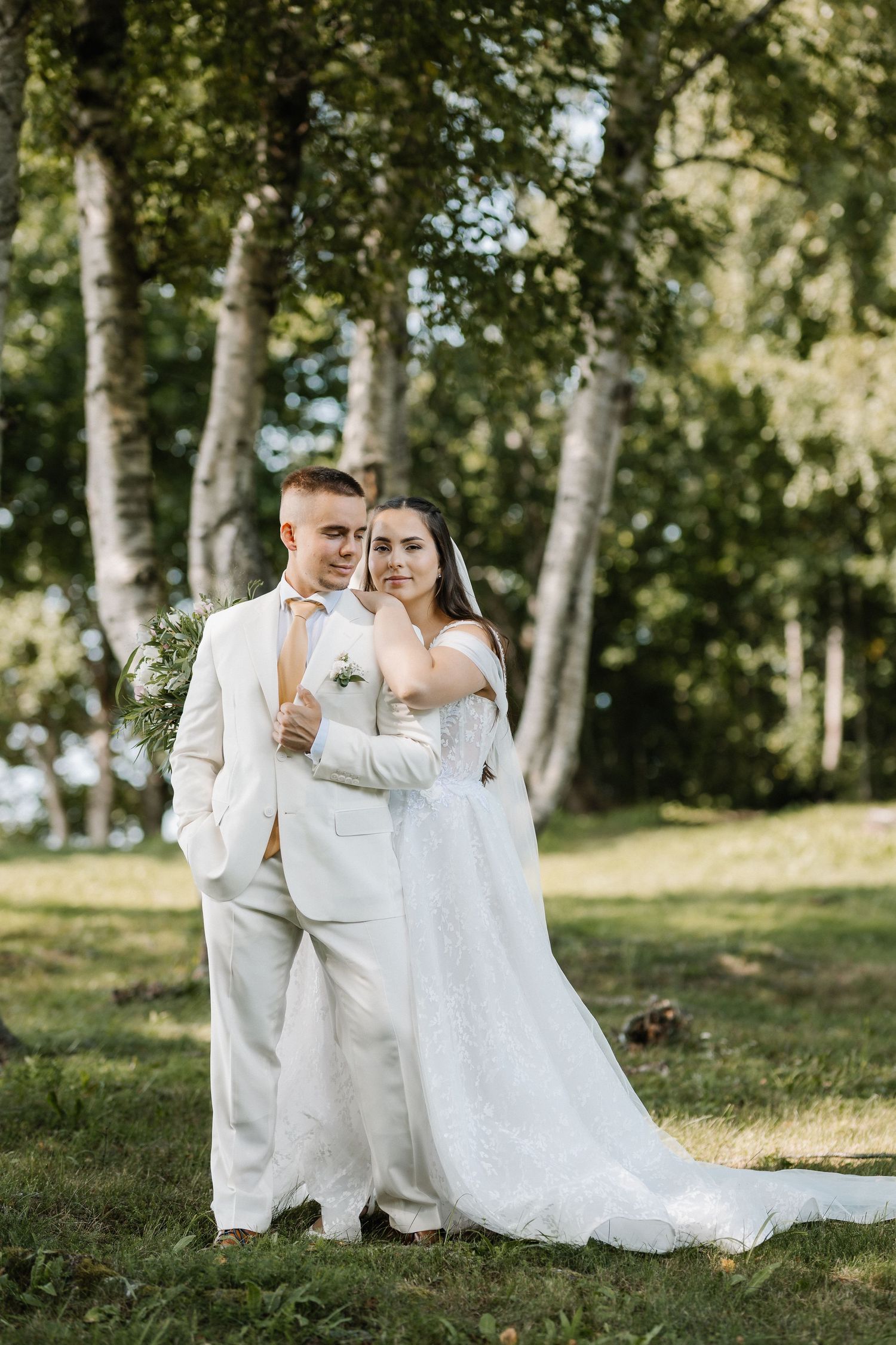 A bride in a white lace gown embraces her groom in a white suit outdoors among birch trees on their wedding day.