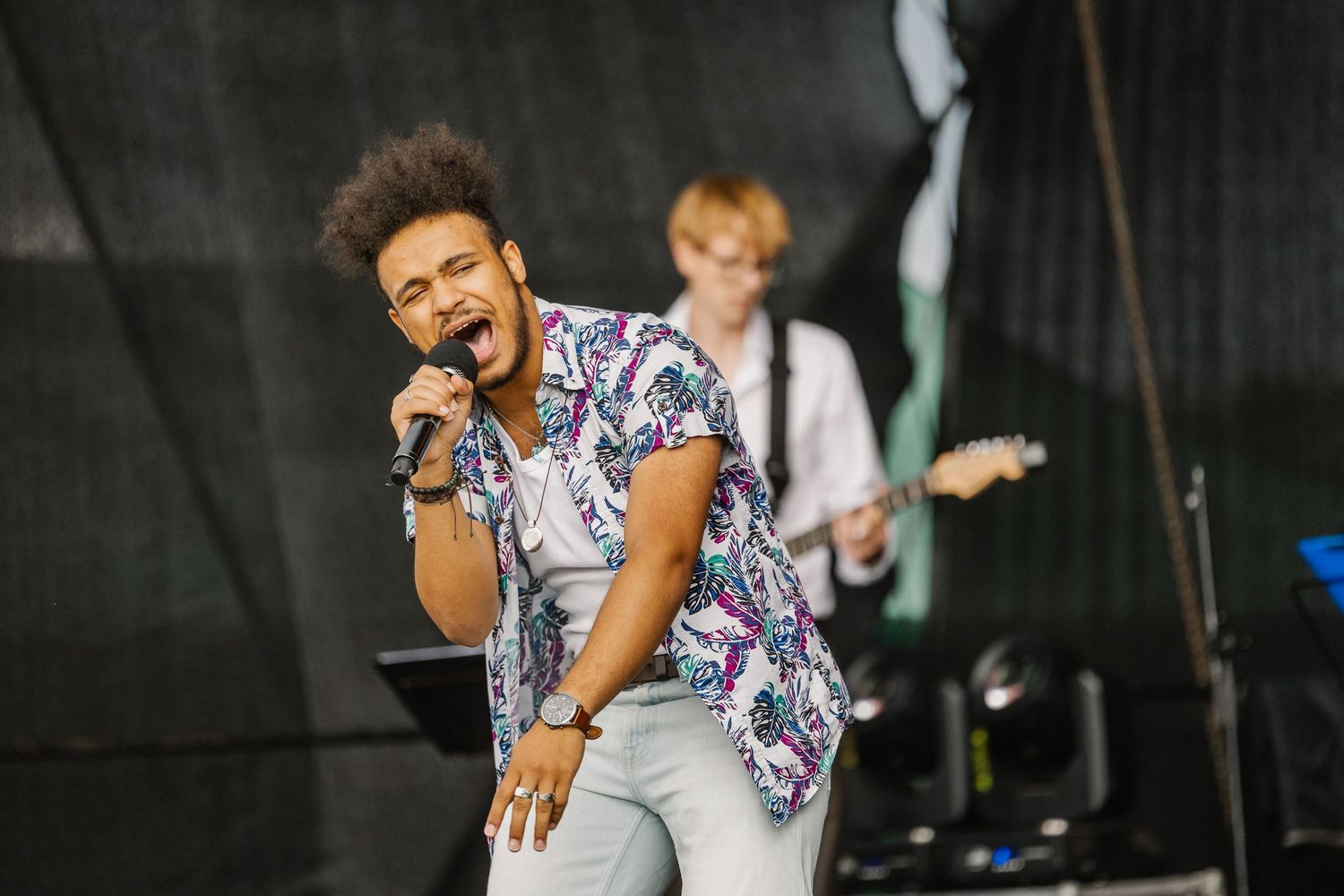 Young male singer performs passionately on stage with microphone, wearing floral shirt, guitarist visible in background.