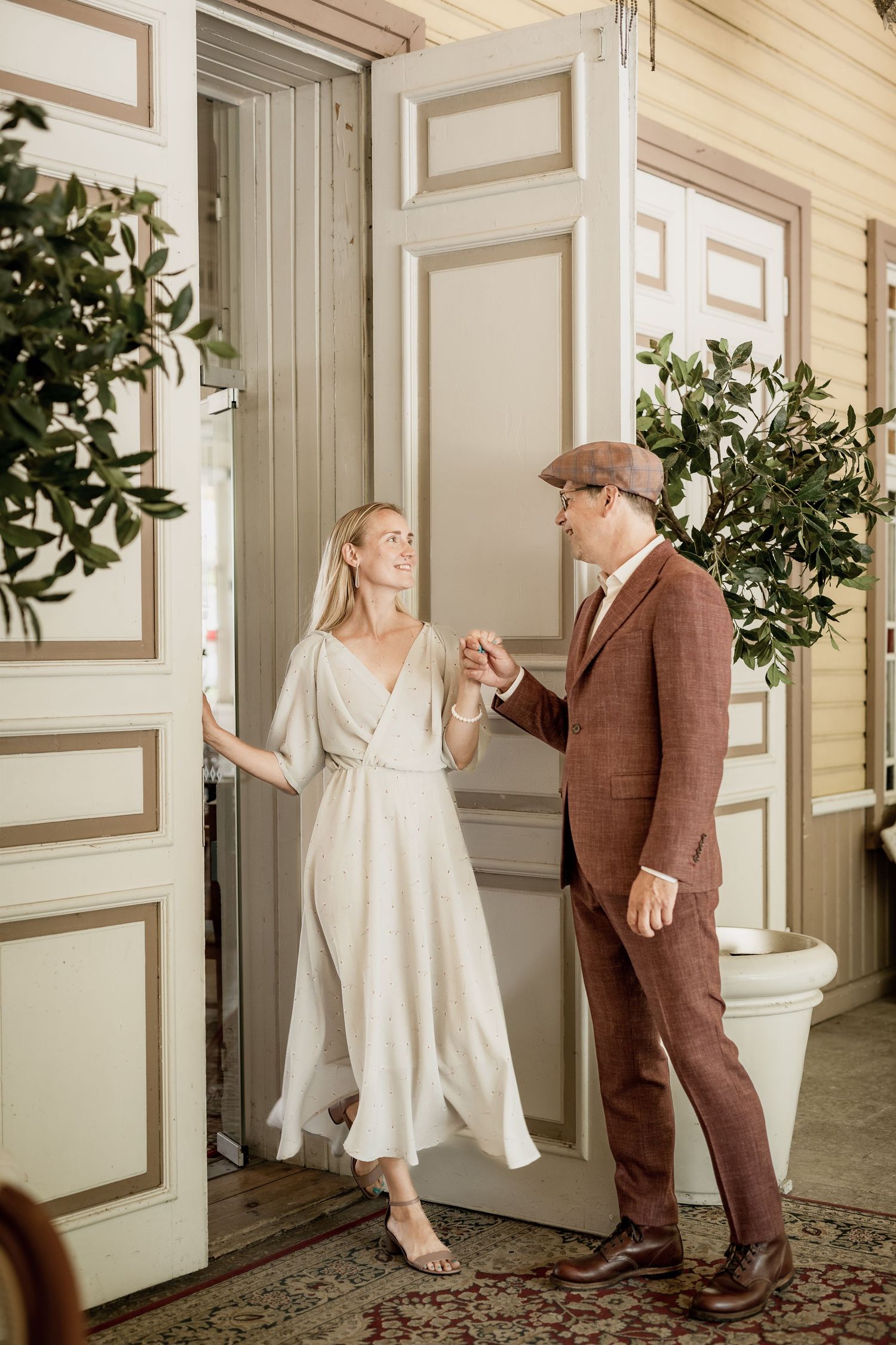 Stylish couple in vintage attire holding hands at elegant white doorway with potted plants nearby.