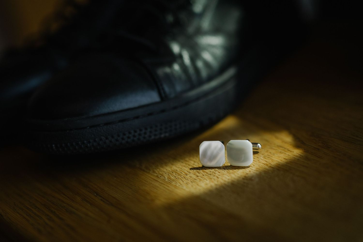 White square cufflinks resting on wooden surface beside a black leather shoe in soft spotlight.