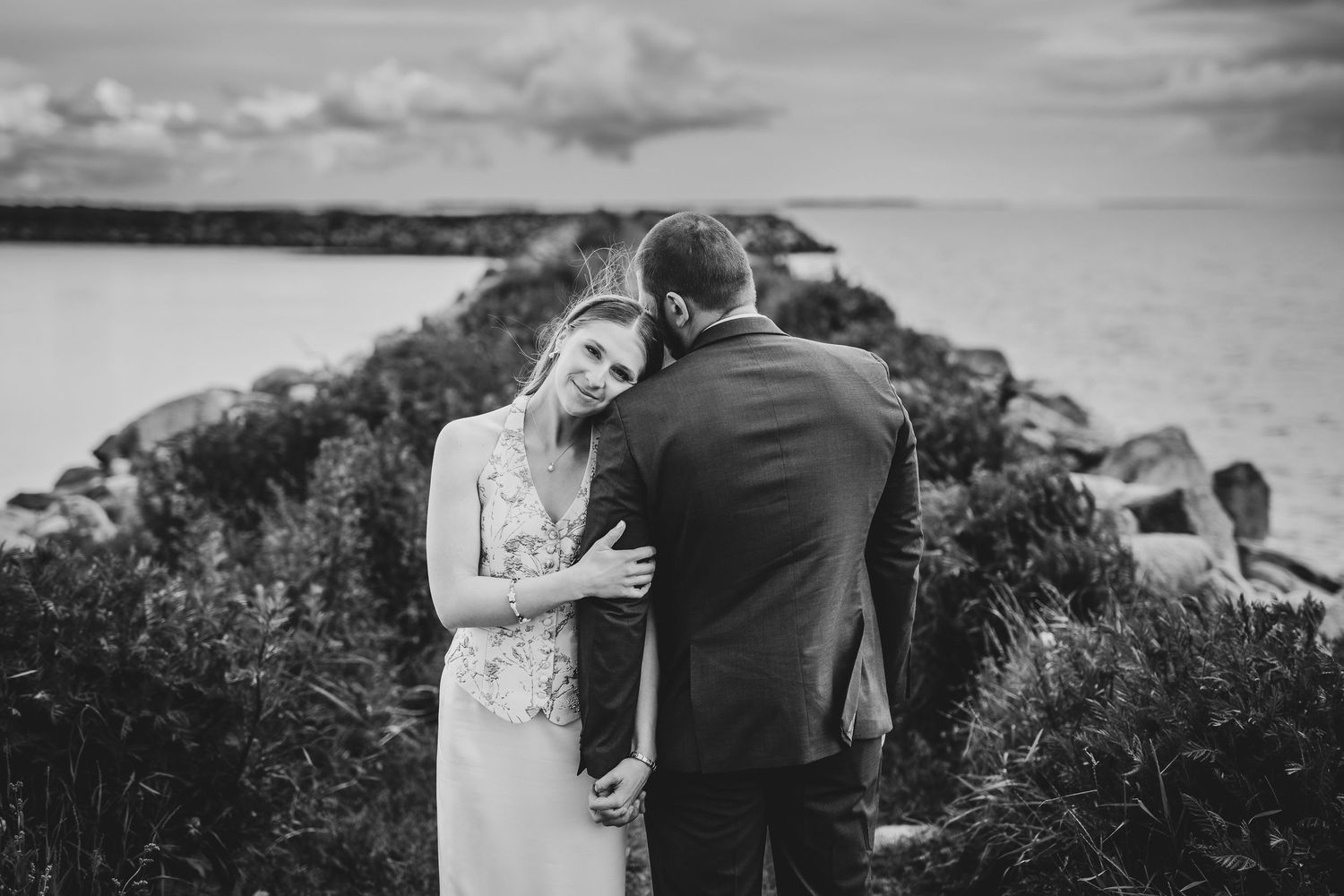 Black and white photo of a bride and groom sharing a tender moment by a rocky seaside, with dramatic cloudy skies.