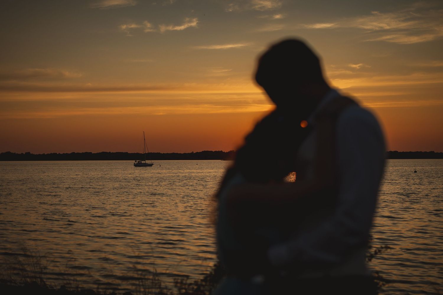 Silhouette of couple embracing at golden sunset over calm water with sailboat in background.