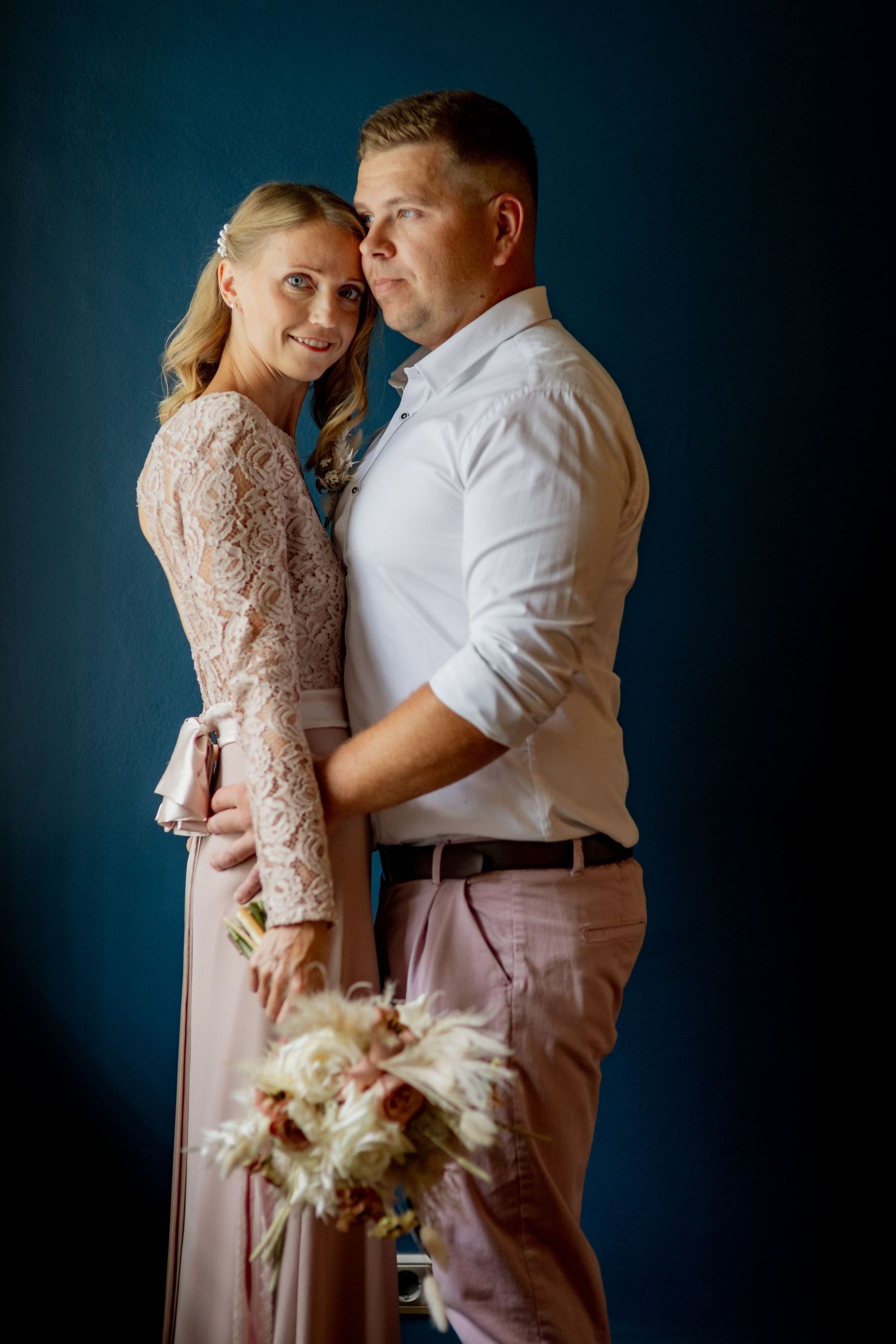 Elegant couple embracing in studio, woman in lace pink dress holding bouquet, man in white shirt, against blue background.
