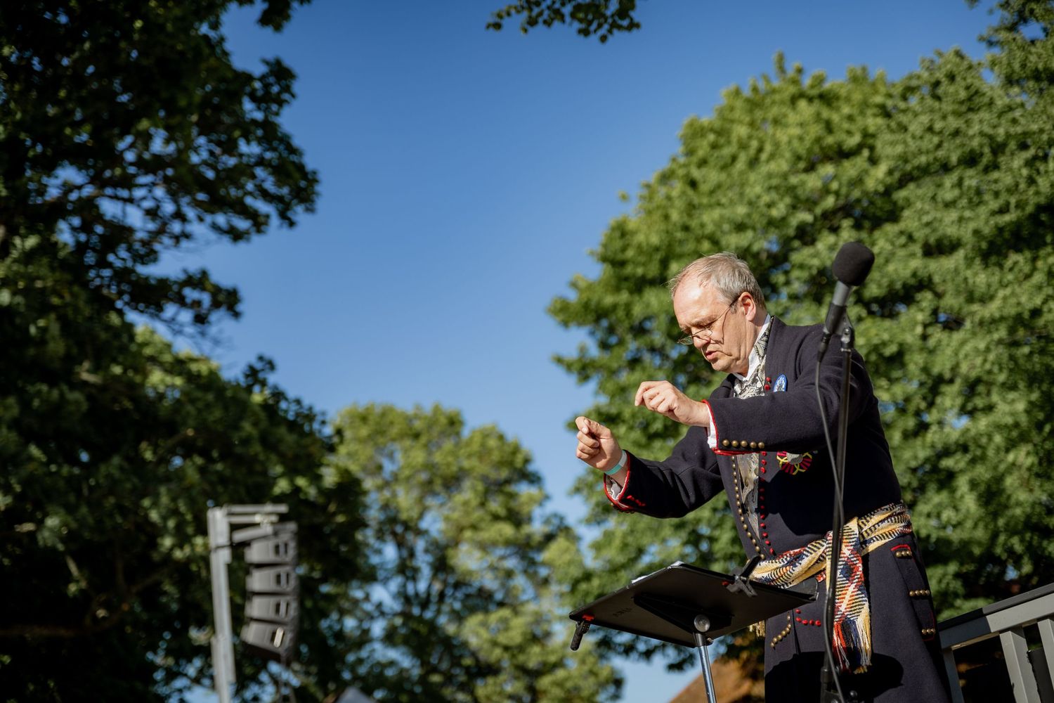 Outdoor conductor leading orchestra under blue sky surrounded by lush green trees in natural amphitheater setting.