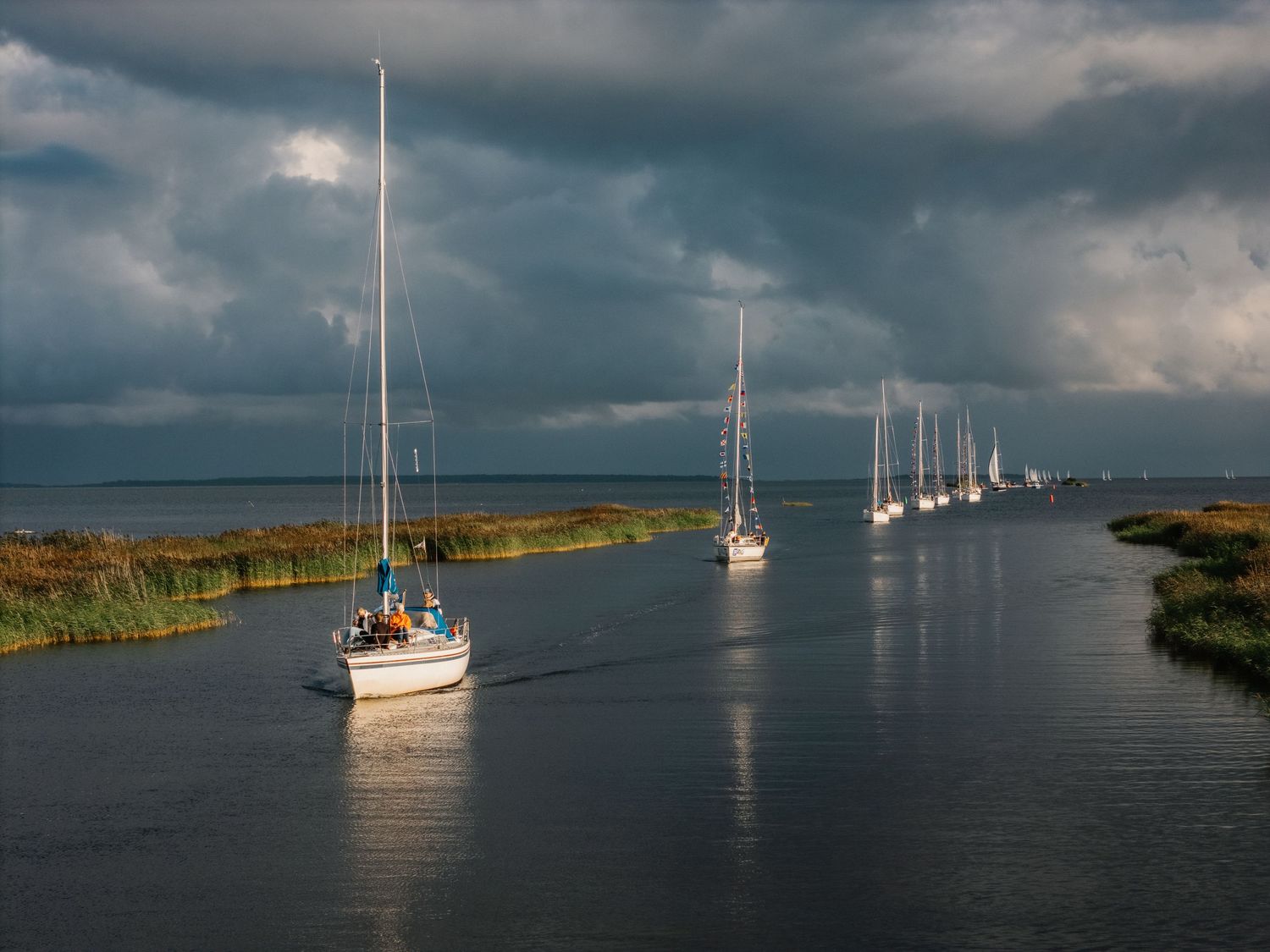 Sailboats navigate a narrow waterway channel under dramatic stormy skies with lush green marshlands on either side.