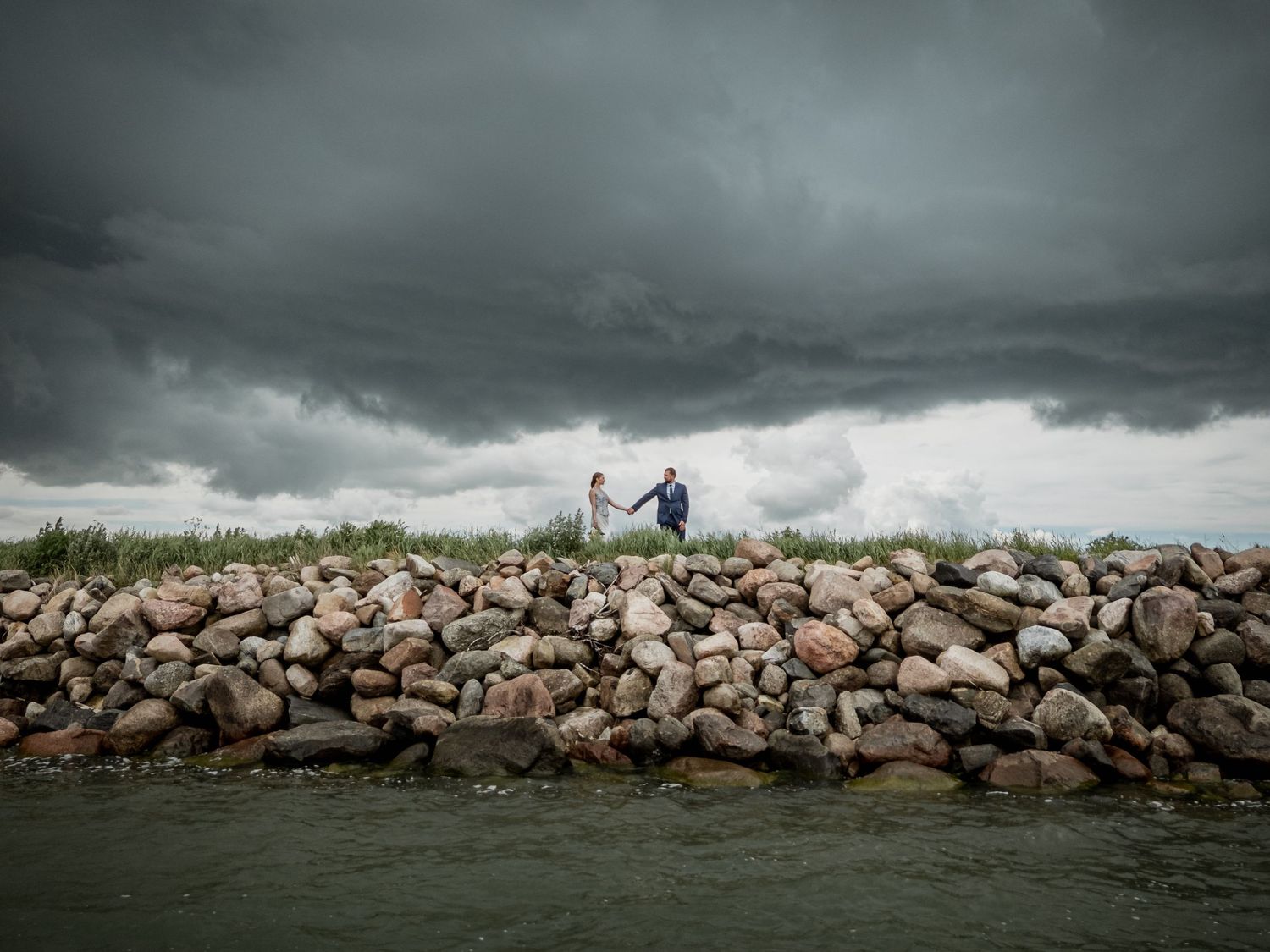 Couple holding hands on rocky jetty under dramatic stormy sky with dark clouds over calm water.