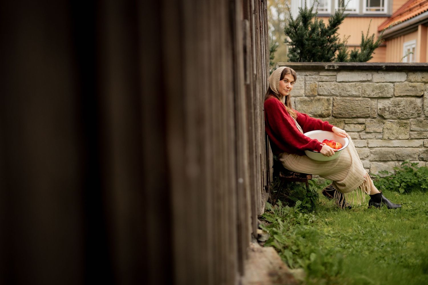 Woman in red sweater sitting against stone wall in garden, relaxing outdoors near wooden fence.